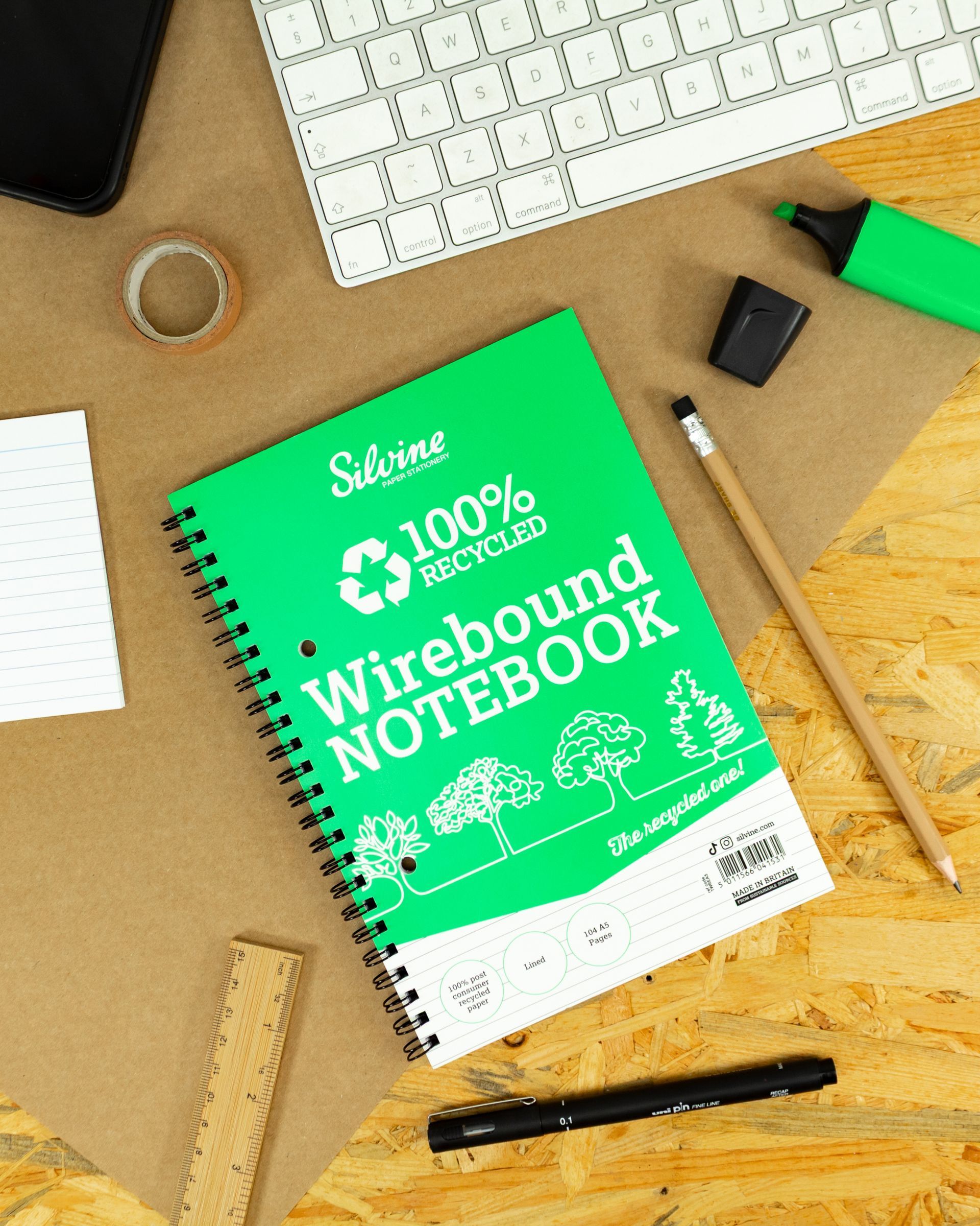 100% recycled notebook on wood desk with keyboard and other stationery items placed around it.