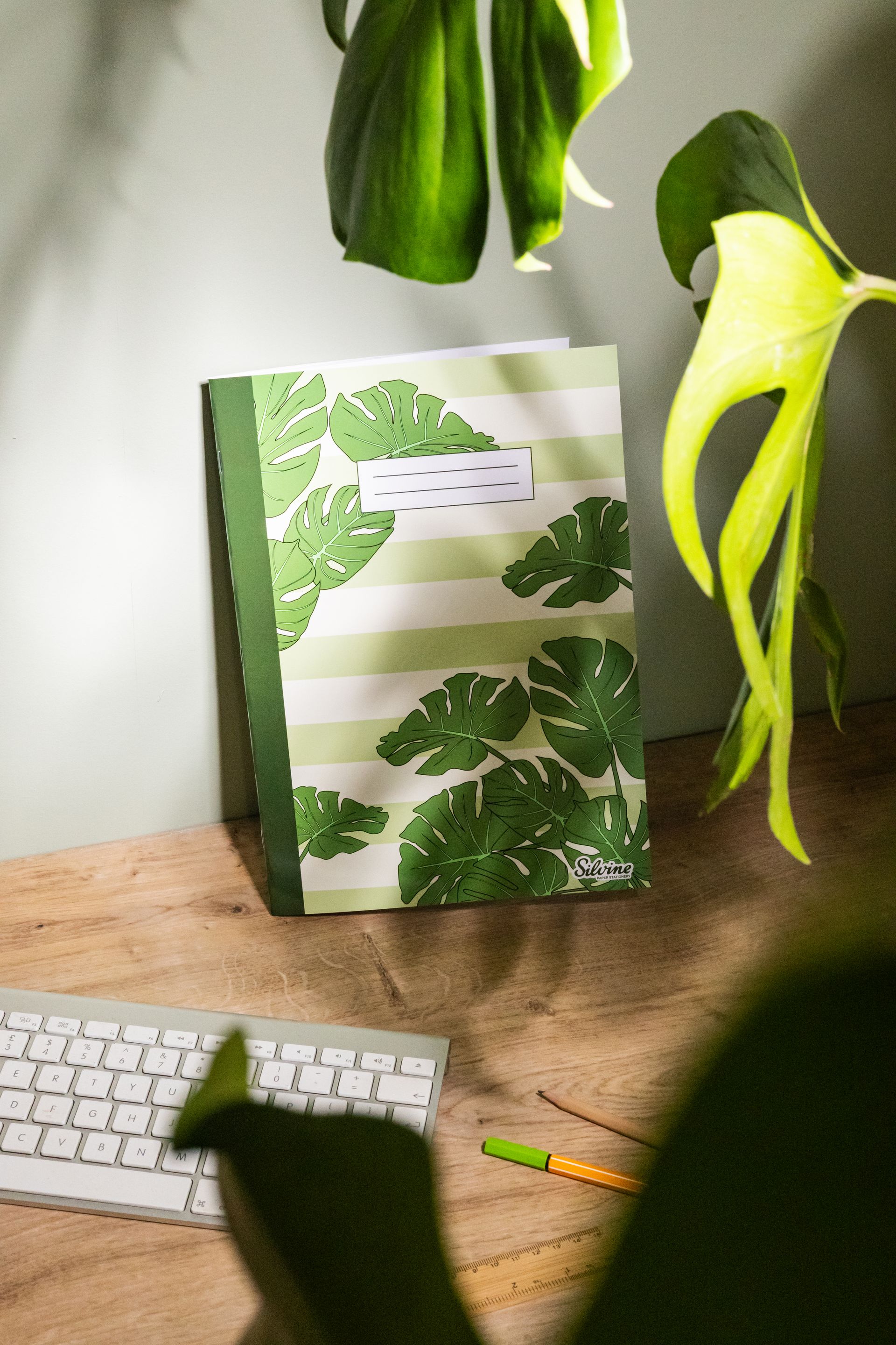Exercise book by Silvine with Monstera Leaf design on the cover. It is on a wooden desk with a Monstera houseplant in view.