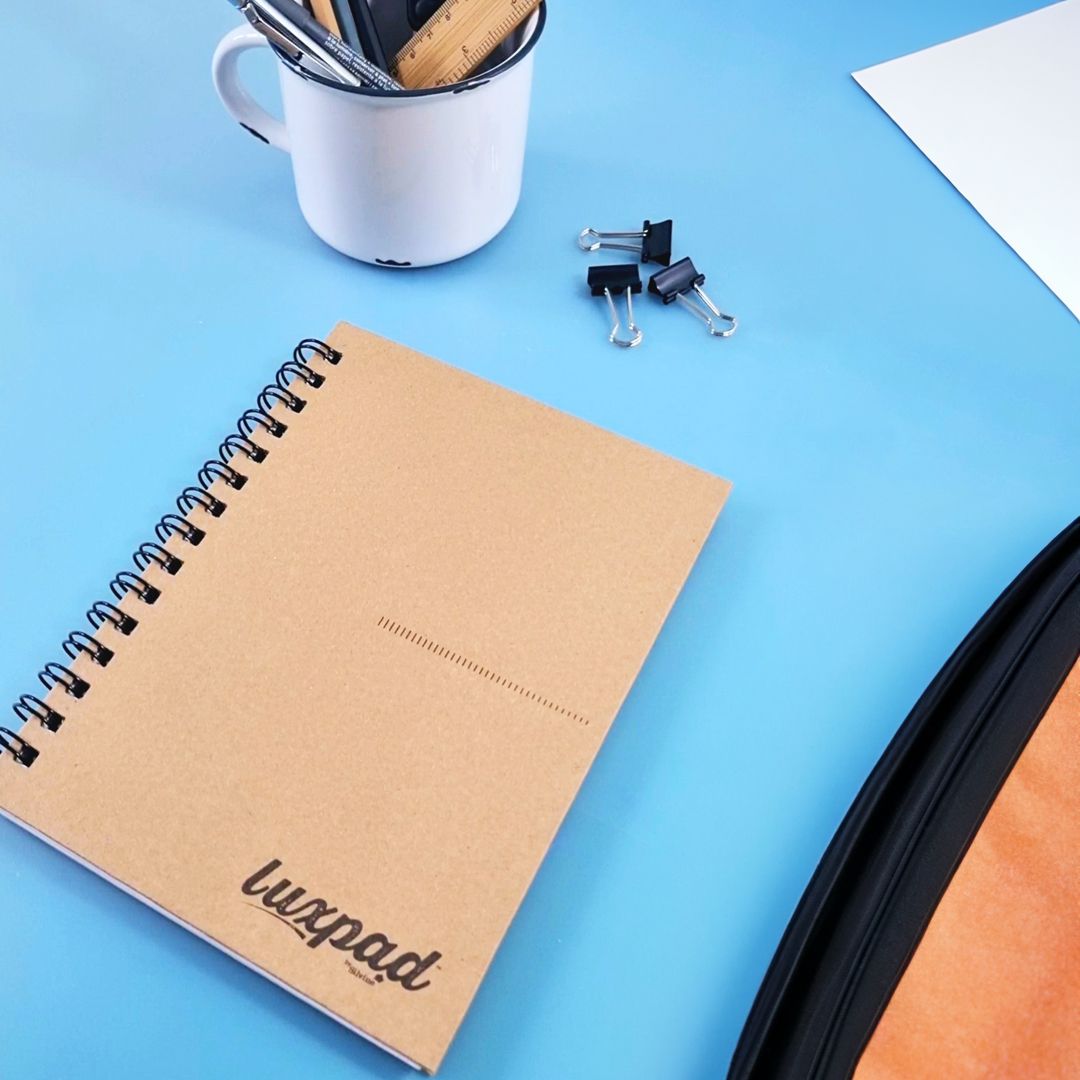 Kraft covered hardback twin wire notebook on blue desk with stationery accessories.