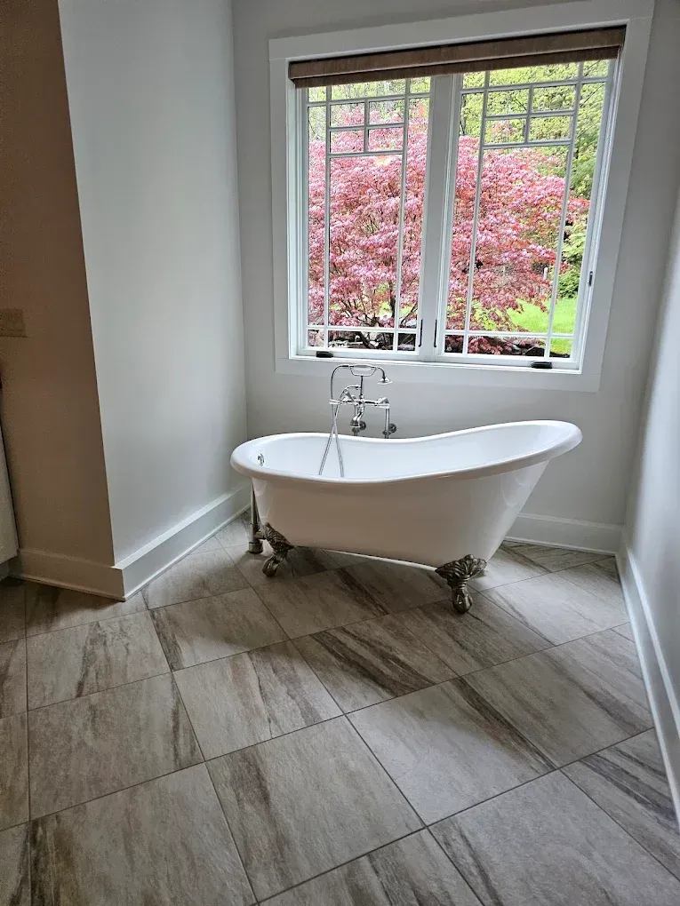 Clawfoot bathtub by a window with a view of pink foliage. Neutral tiled floor and white walls.