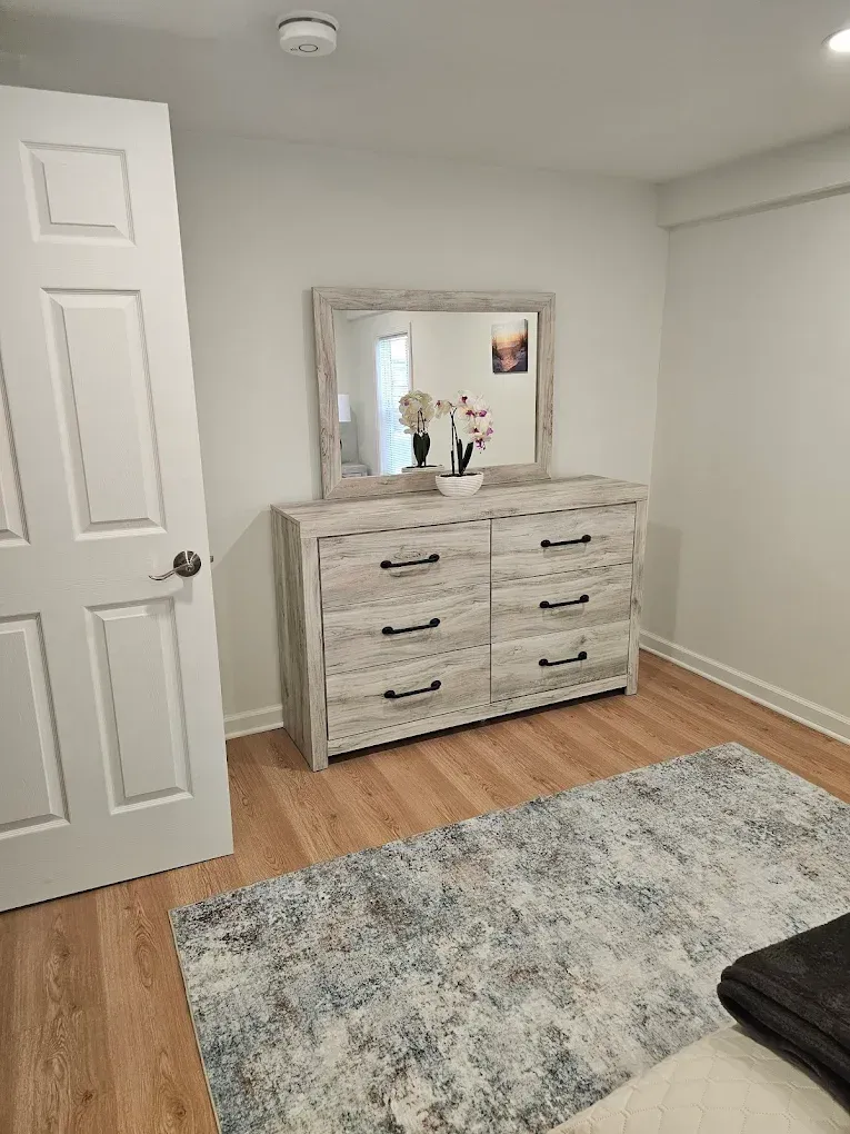 Bedroom with a light wood dresser, mirror, and rug on a wood floor. A door is on the left.