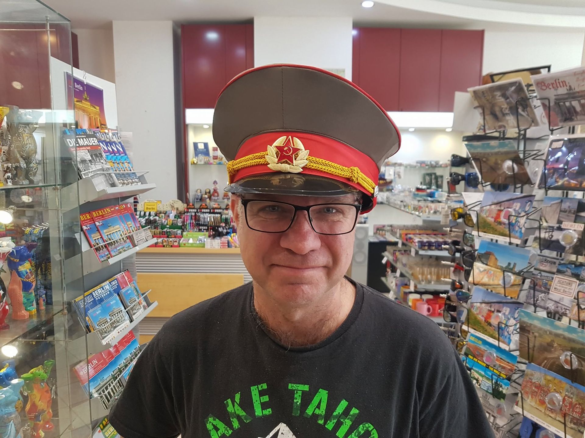 Een man staat lachend in een speelgoedwinkel, met een grote rode pet en een zwart T-shirt aan.