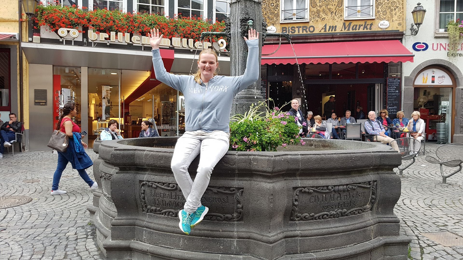Een persoon in een licht shirt en een witte broek springt op een stenen fontein op een druk plein met winkels en bloemen in de buurt.