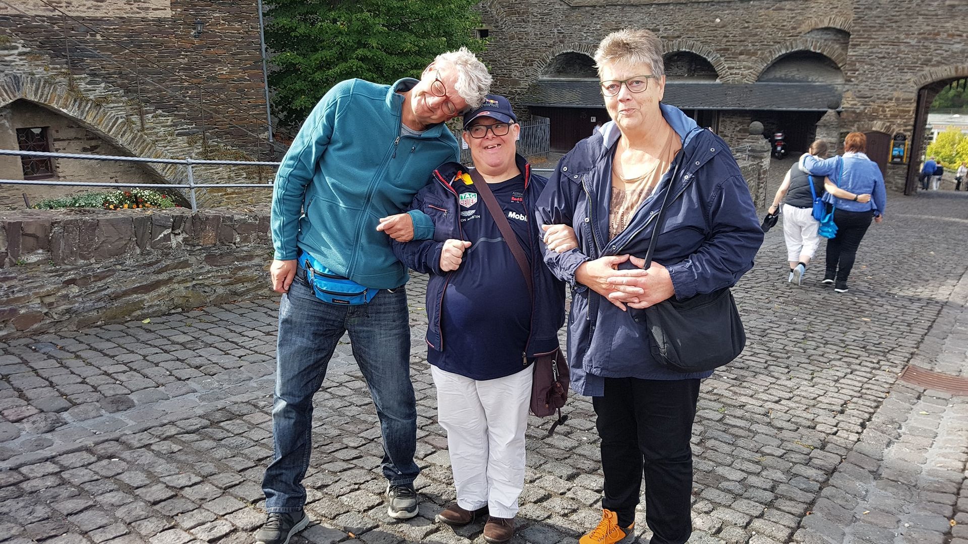 Drie mensen poseren samen op een straat met kinderkopjes voor een oude bakstenen muur.