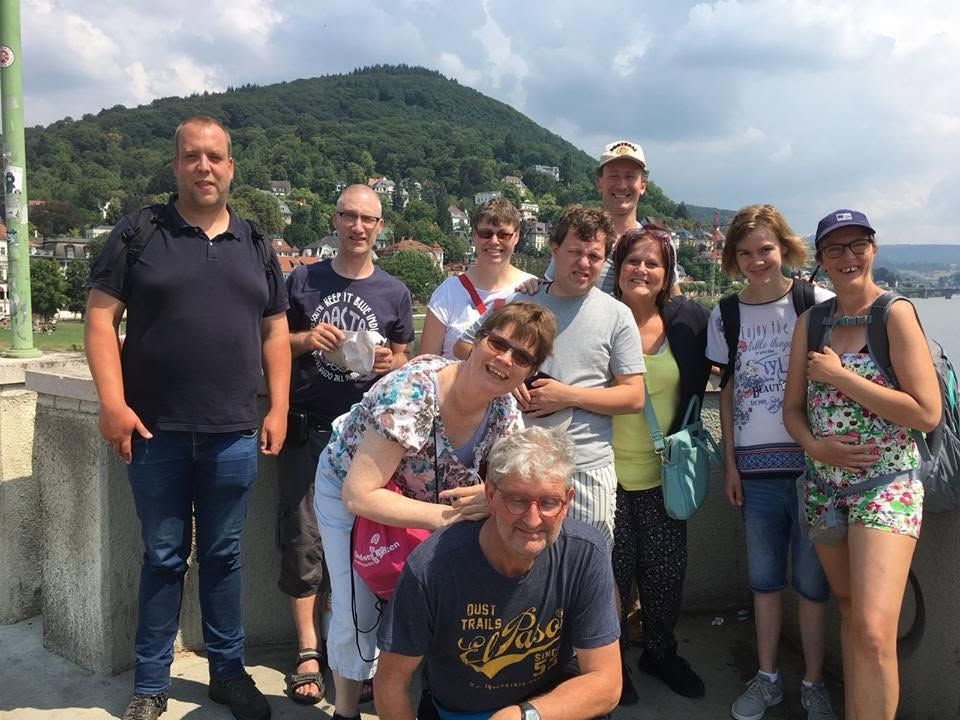 Een groep mensen poseert buiten op een zonnige heuvel met bergen en water op de achtergrond.