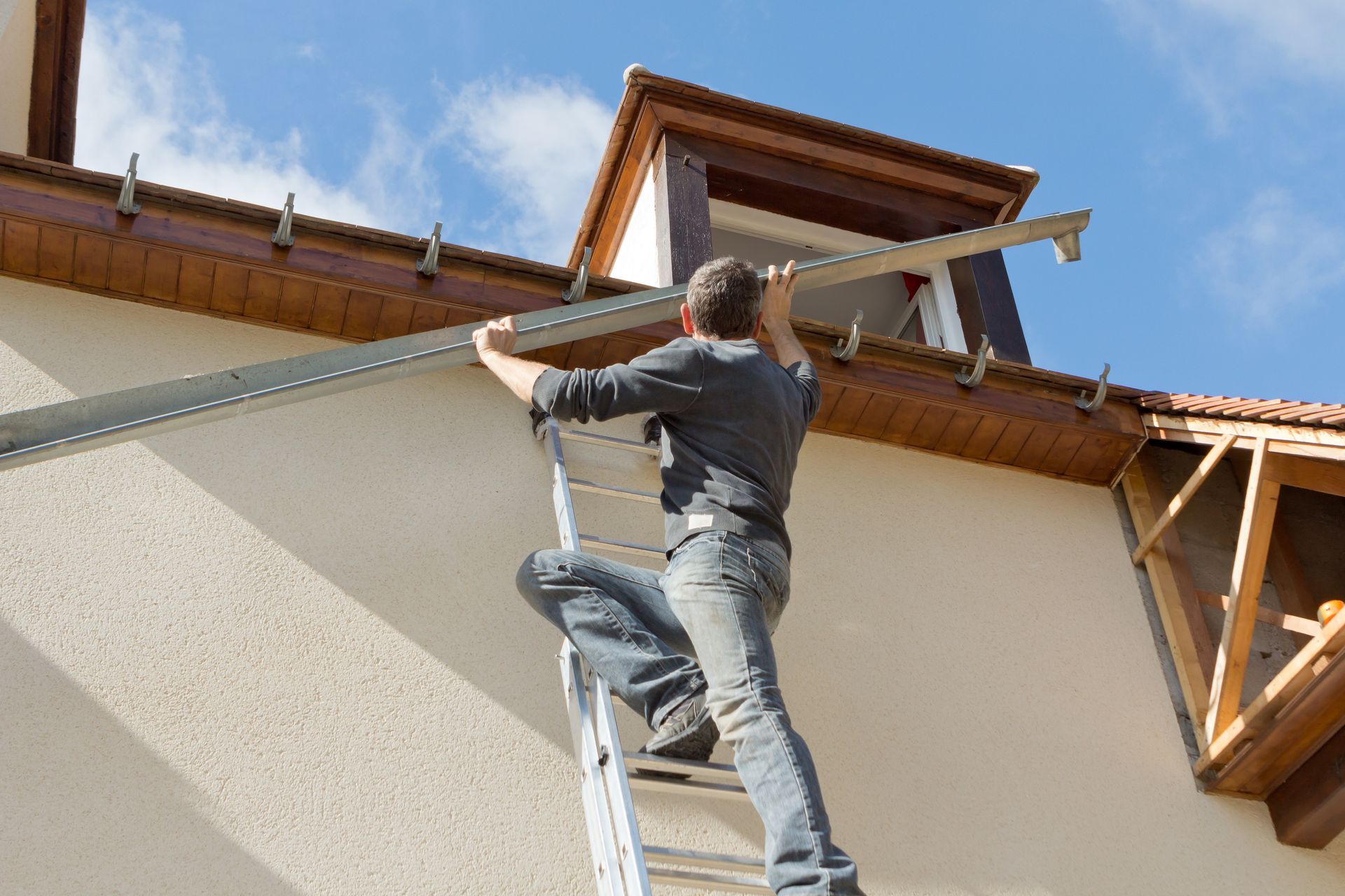 A person on a ladder installs a metal gutter section along the eave of a house against a blue sky.