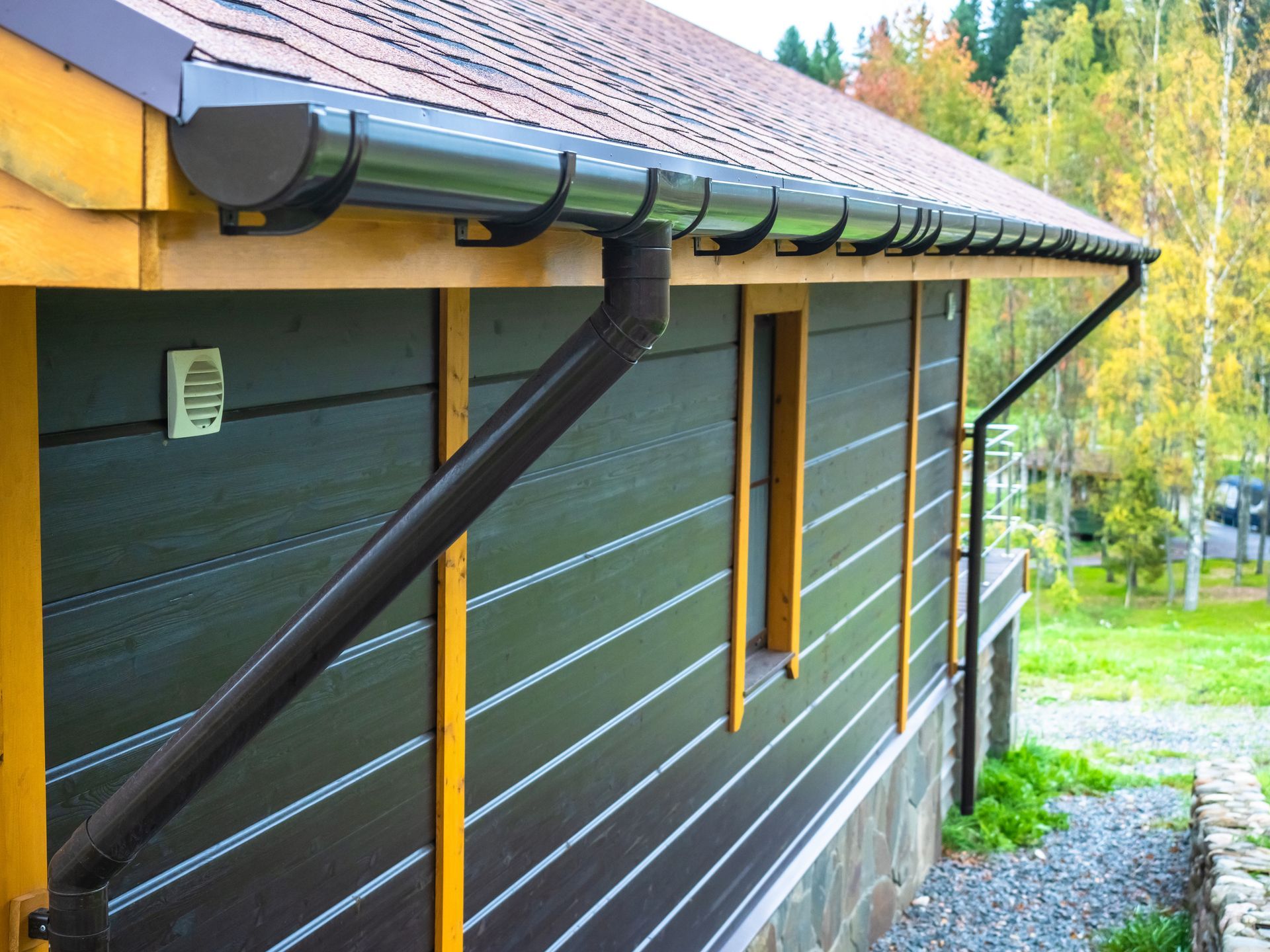A dark brown gutter system with a downspout installed on the side of a modern house with wood-paneled walls.