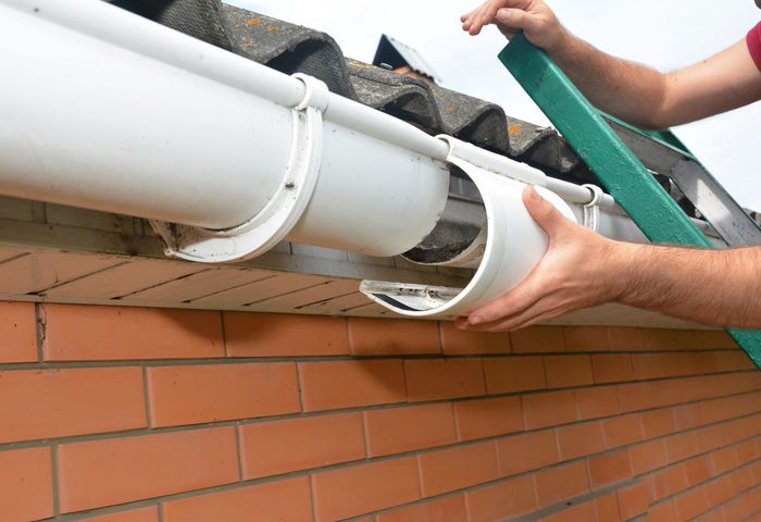 A person on a ladder repairs a white plastic rain gutter attached to a brick building.
