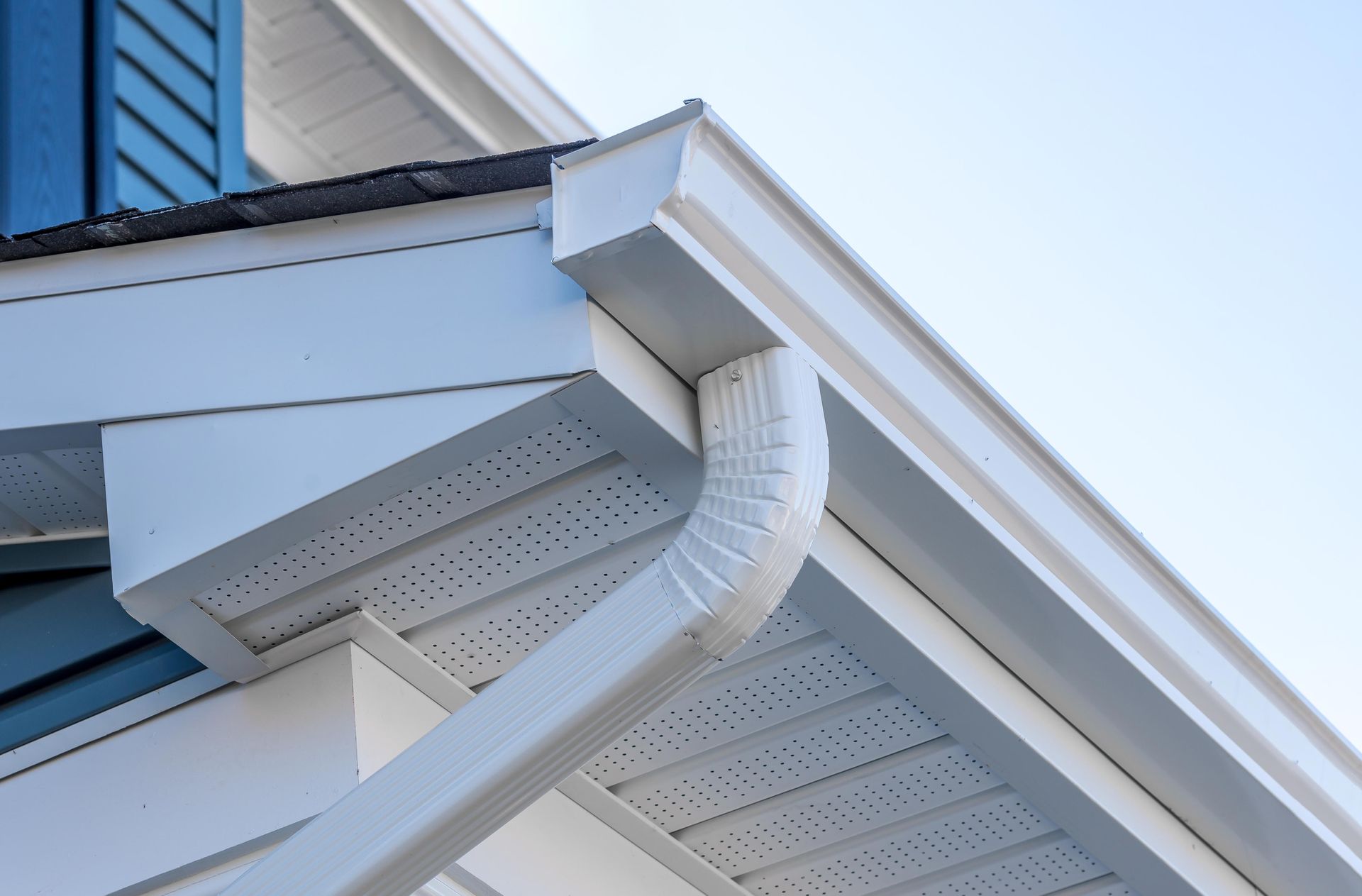White rain gutters and a downspout attached to the white eaves and roofline of a blue-sided house.