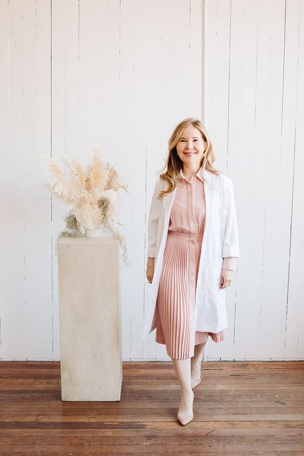 A woman in a pink dress and white coat is standing next to a vase of flowers.