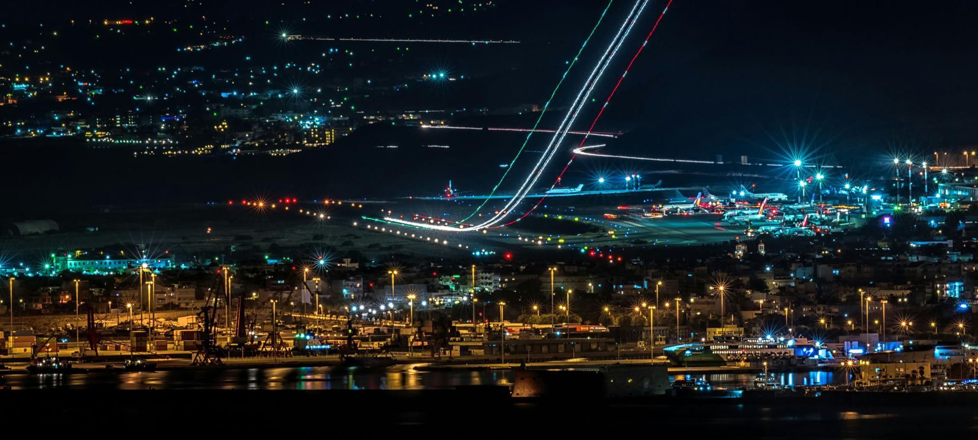Night view of city lights and a descending airplane's light trails over an airport.