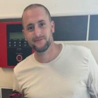 Man with a light beard smiles, wearing a cream shirt. Leans near red panel with buttons.