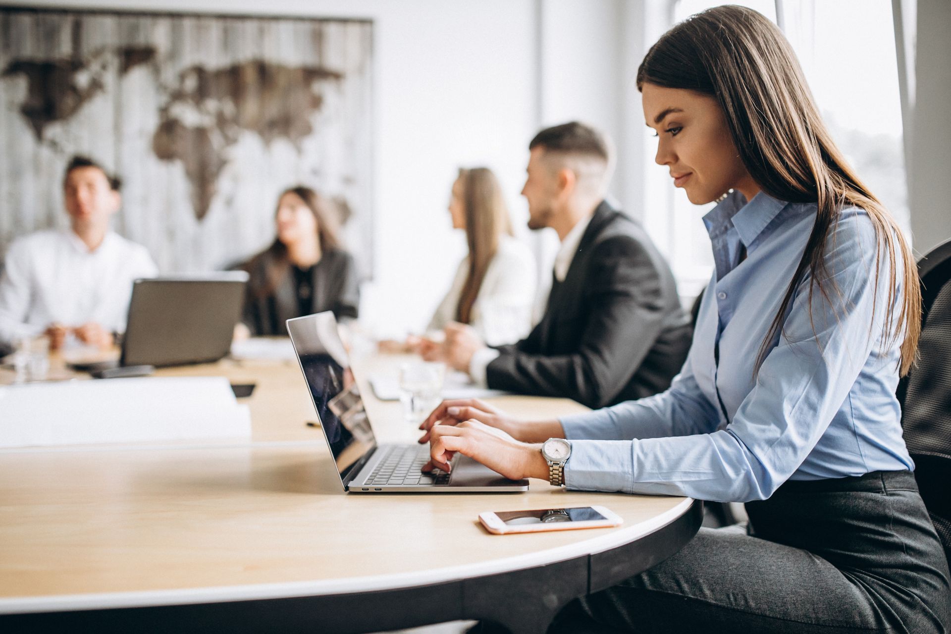 Woman typing on laptop at a table, people in background at meeting, office setting.