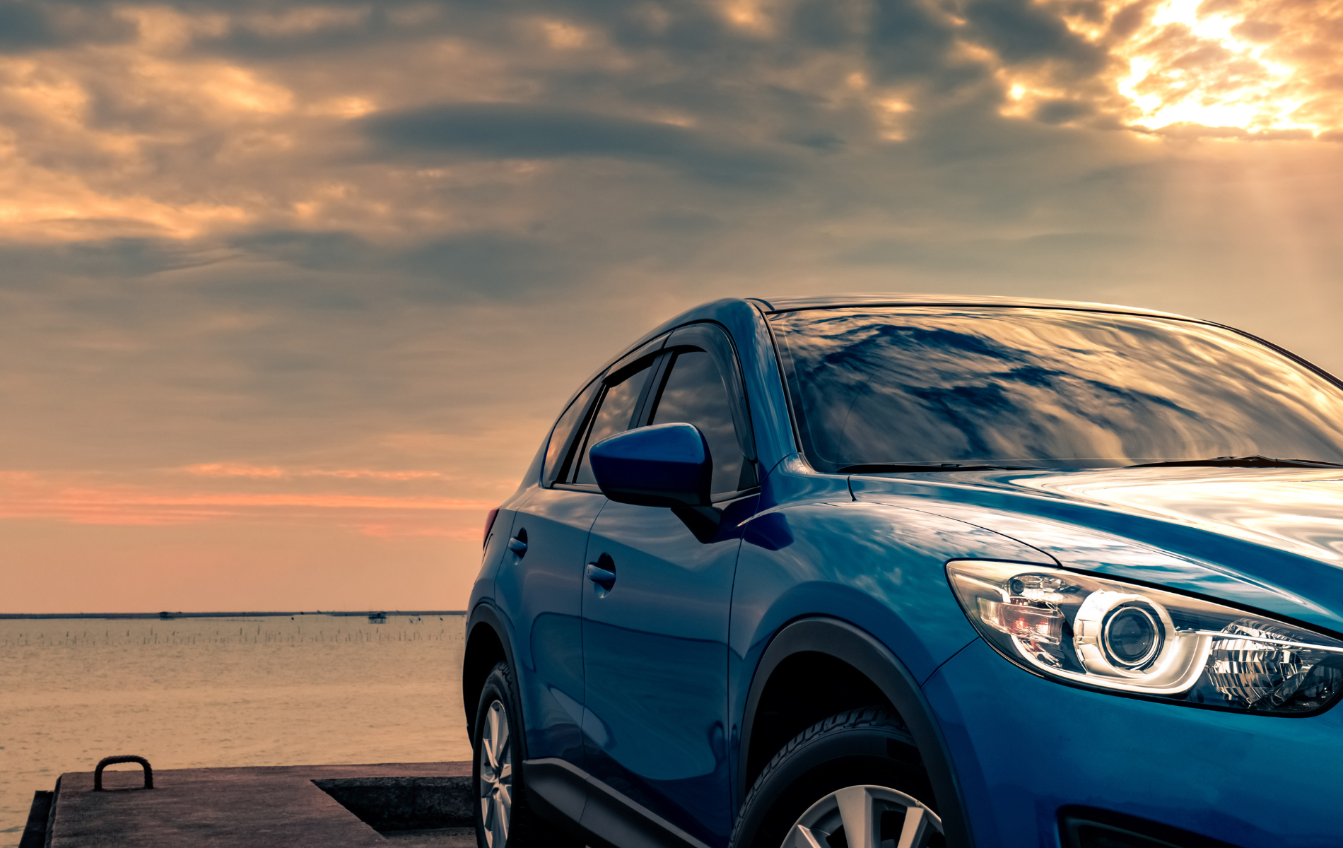 A blue car is parked on a dock next to the ocean at sunset.