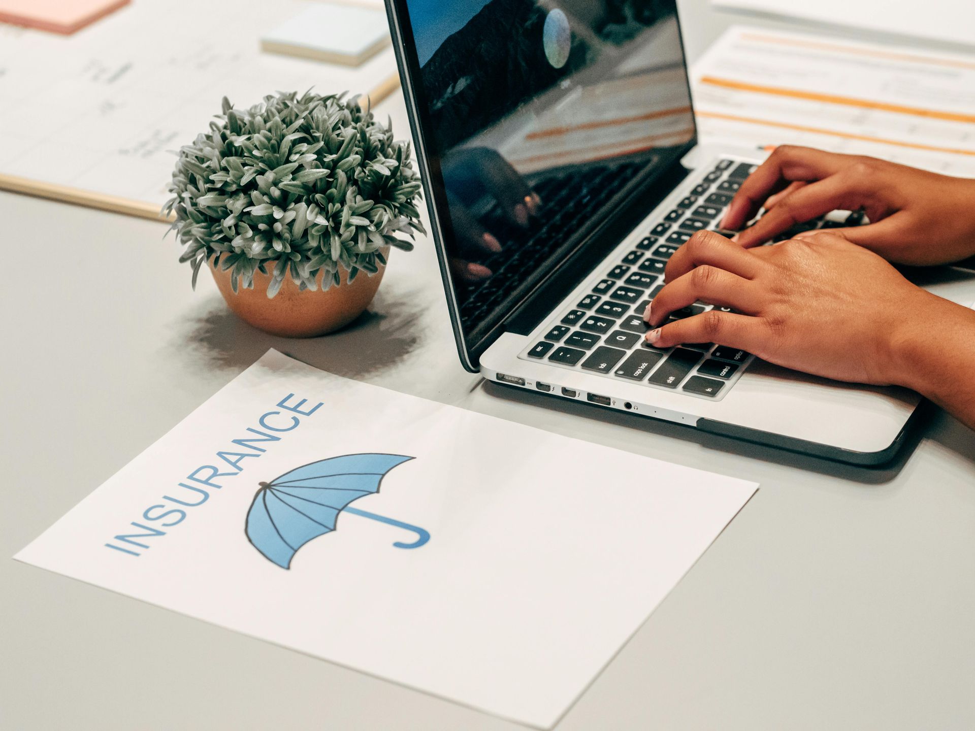 Hands typing on laptop beside a paper labeled “Insurance” with an umbrella icon and small potted plant.