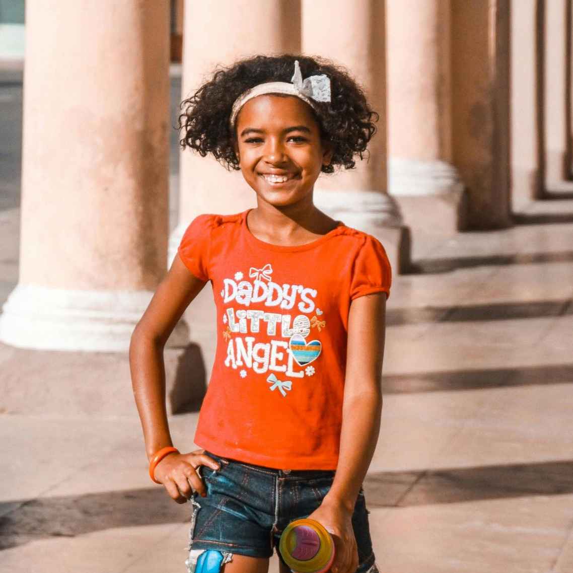 Una chica con una camisa naranja y pantalones cortos de mezclilla sonríe, parada frente a unas columnas.