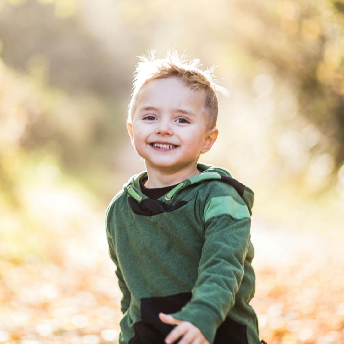Niño pequeño sonriendo con una sudadera con capucha verde al aire libre, con follaje otoñal de fondo.