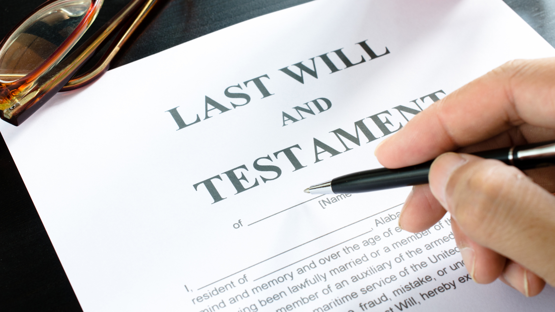 Hand with pen on a Last Will and Testament document, glasses in the corner.