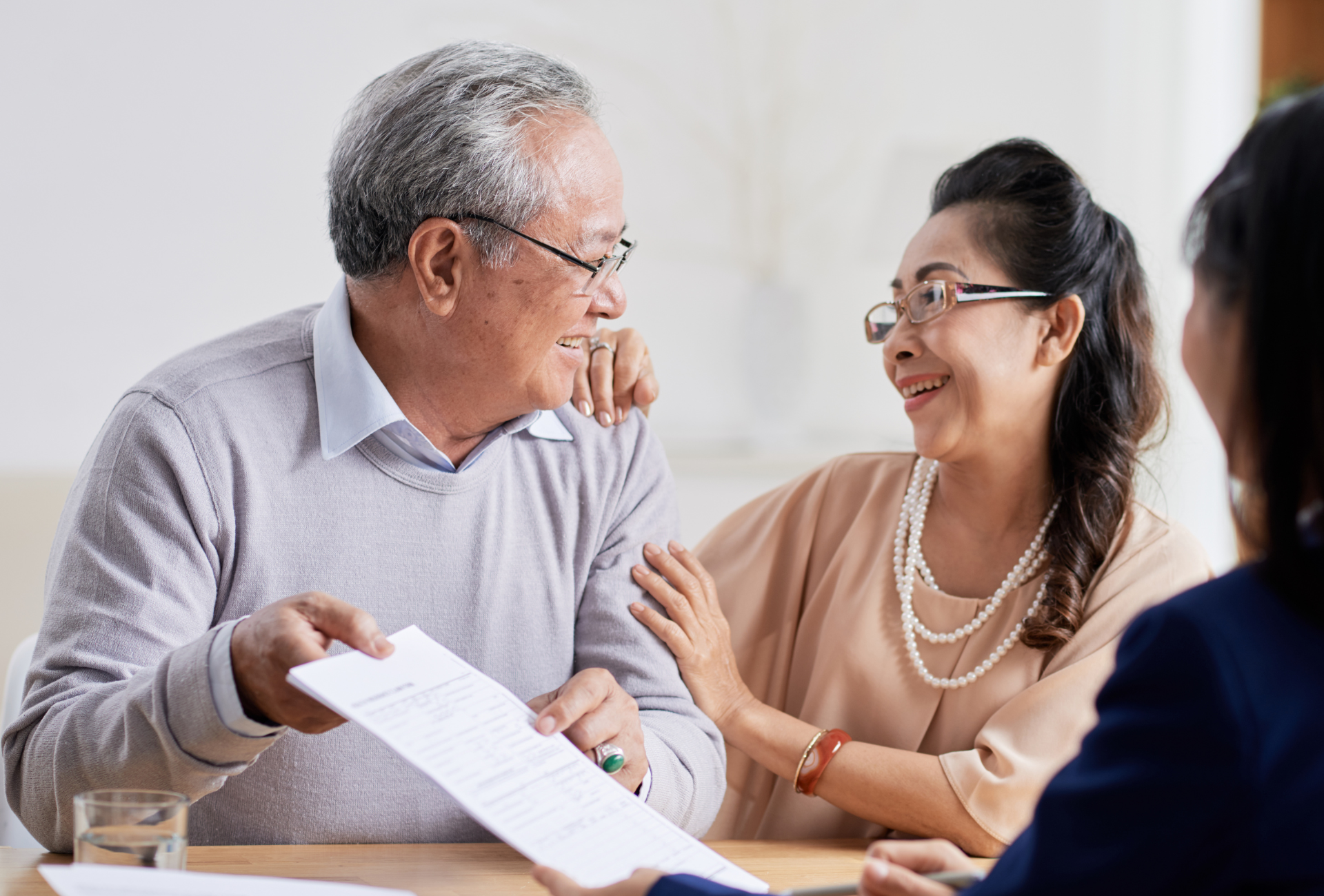 An elderly couple is sitting at a table talking to a woman while holding a piece of paper.