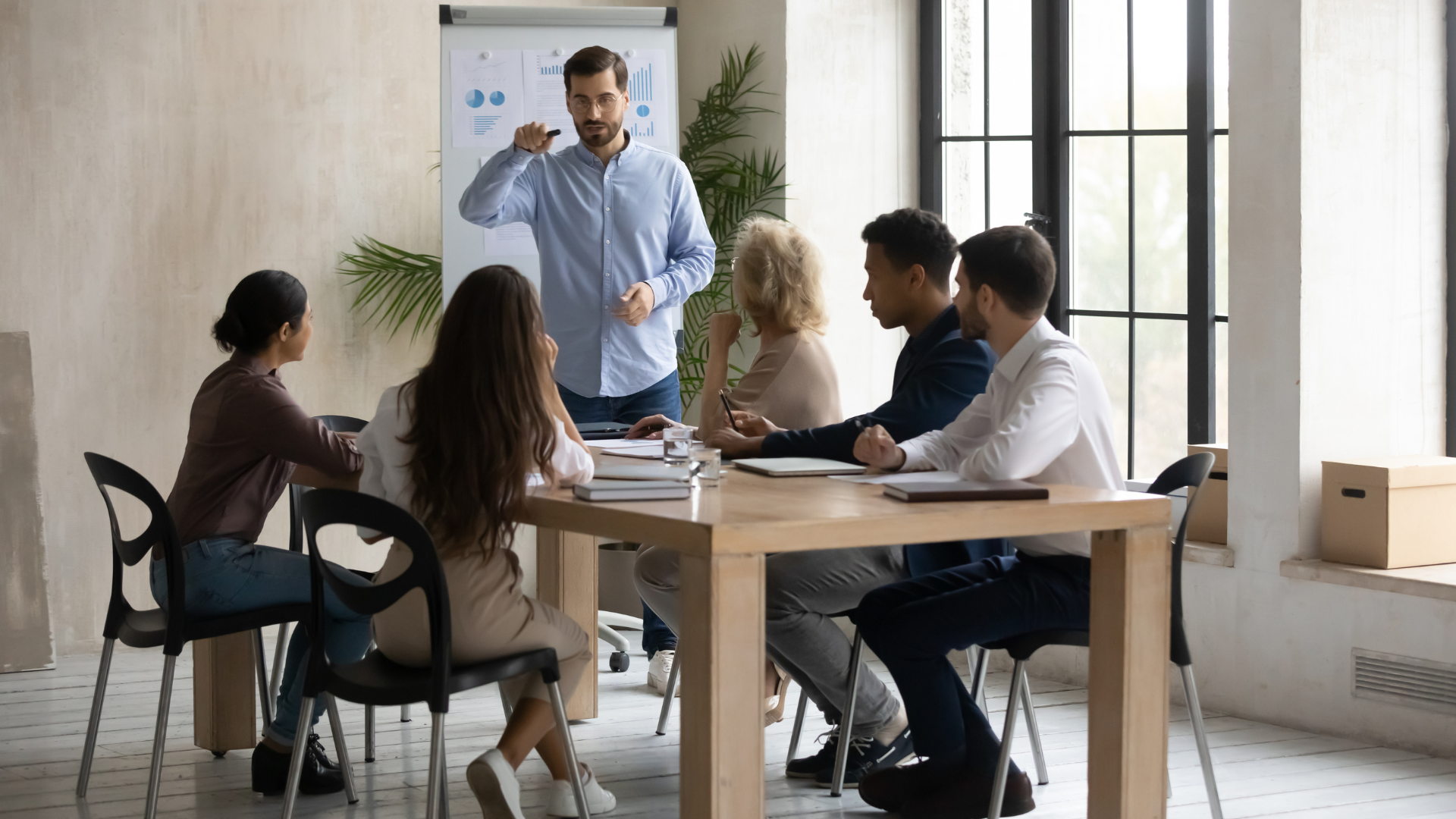 Man leading a meeting, gesturing at a whiteboard. Others sit around a table in an office.