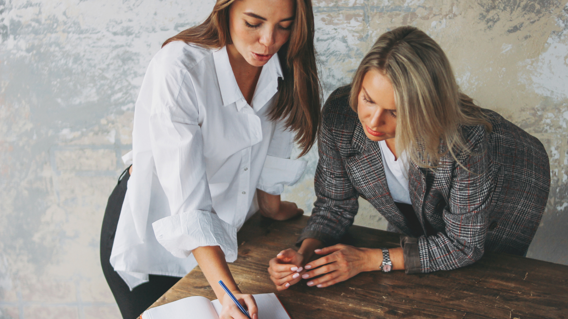 Two women at a desk, reviewing papers, one writes with a pen.