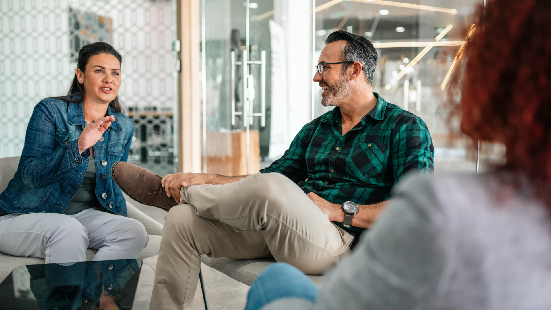 Three people in a modern office, engaged in conversation. Woman gestures, man smiles, other person in the foreground.