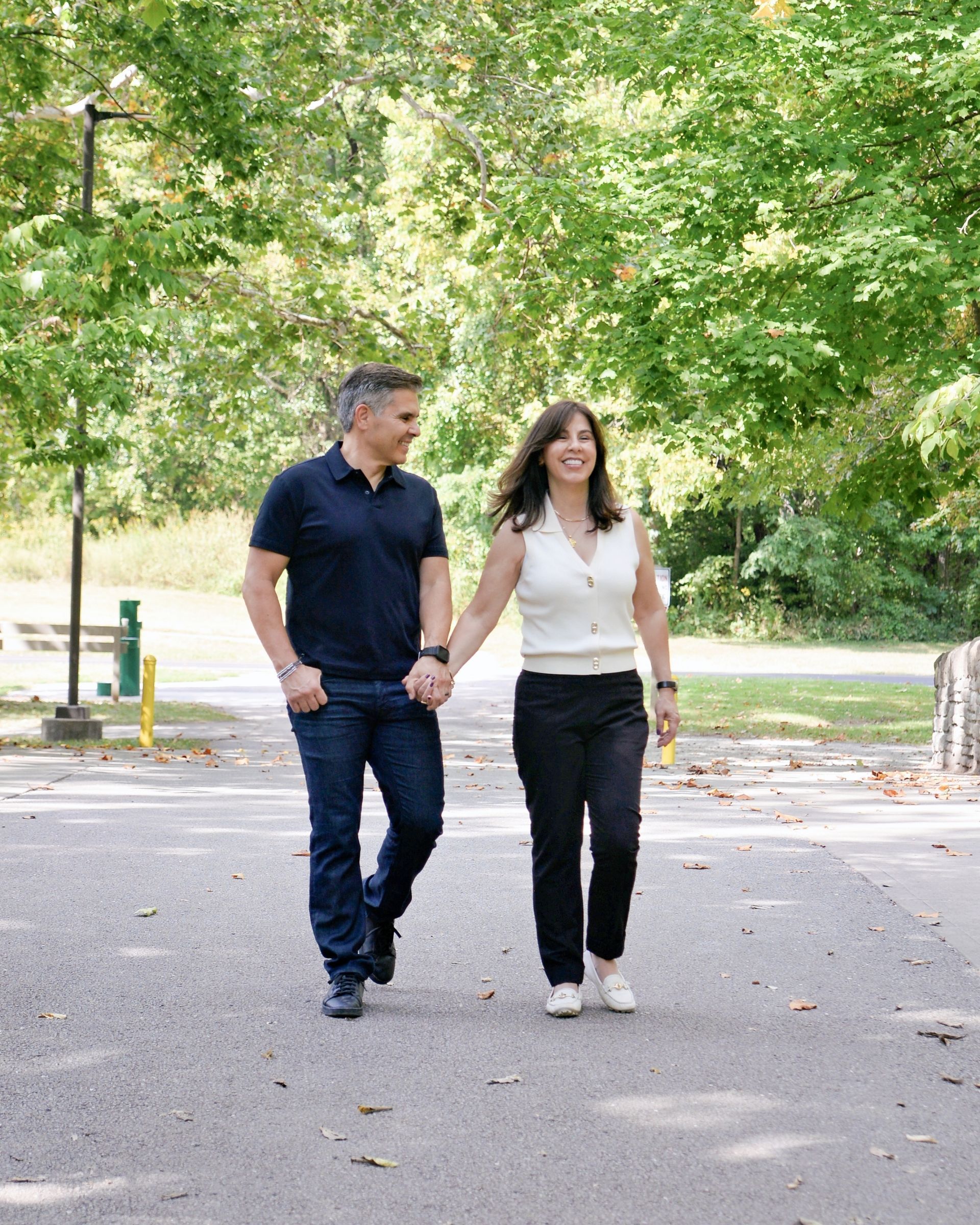 Couple holding hands, walking on a path in a park. Man in blue shirt and jeans, woman in white top and black pants.