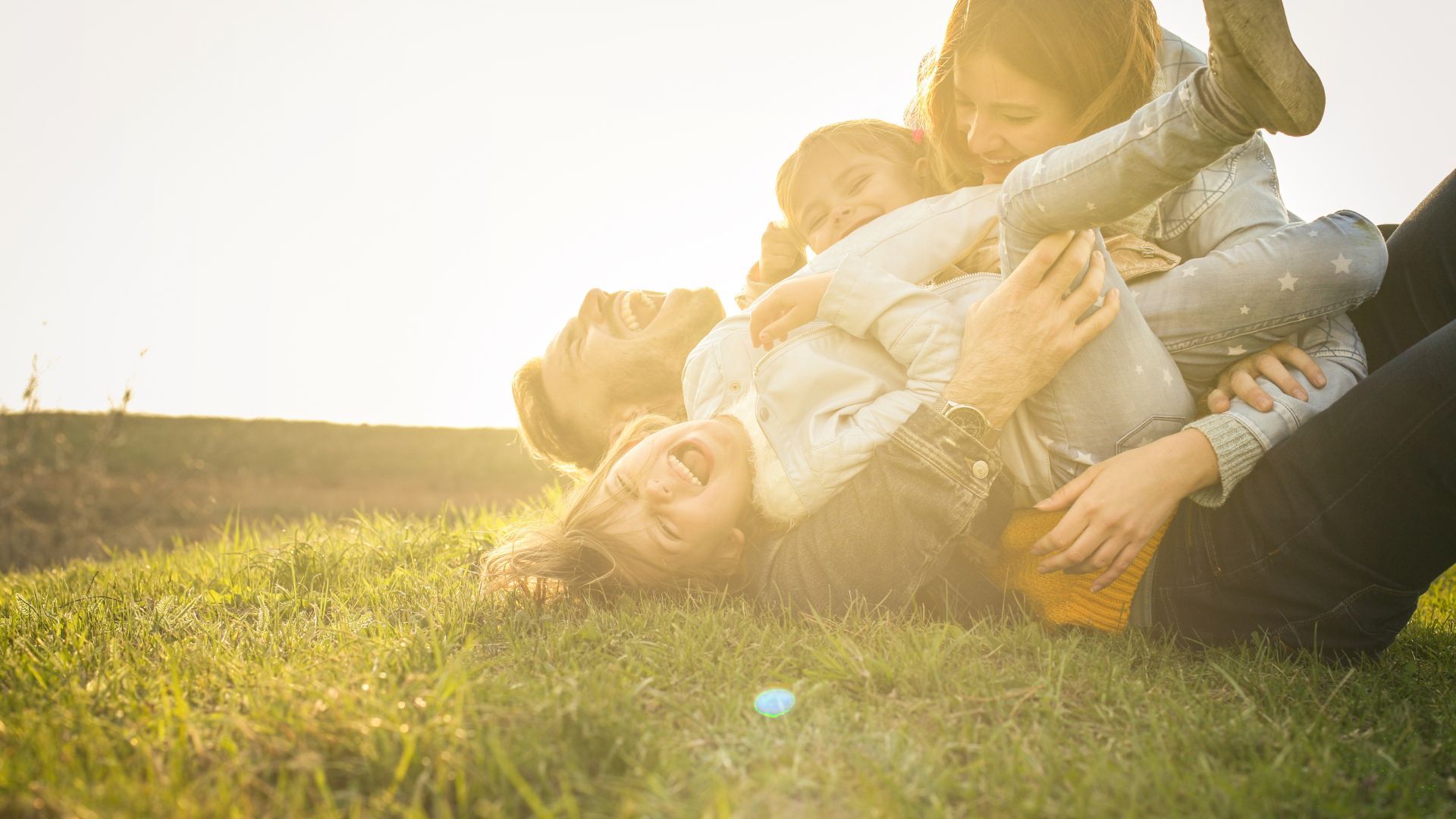 Family of four laughing and playing on a grassy hill, bathed in sunlight.