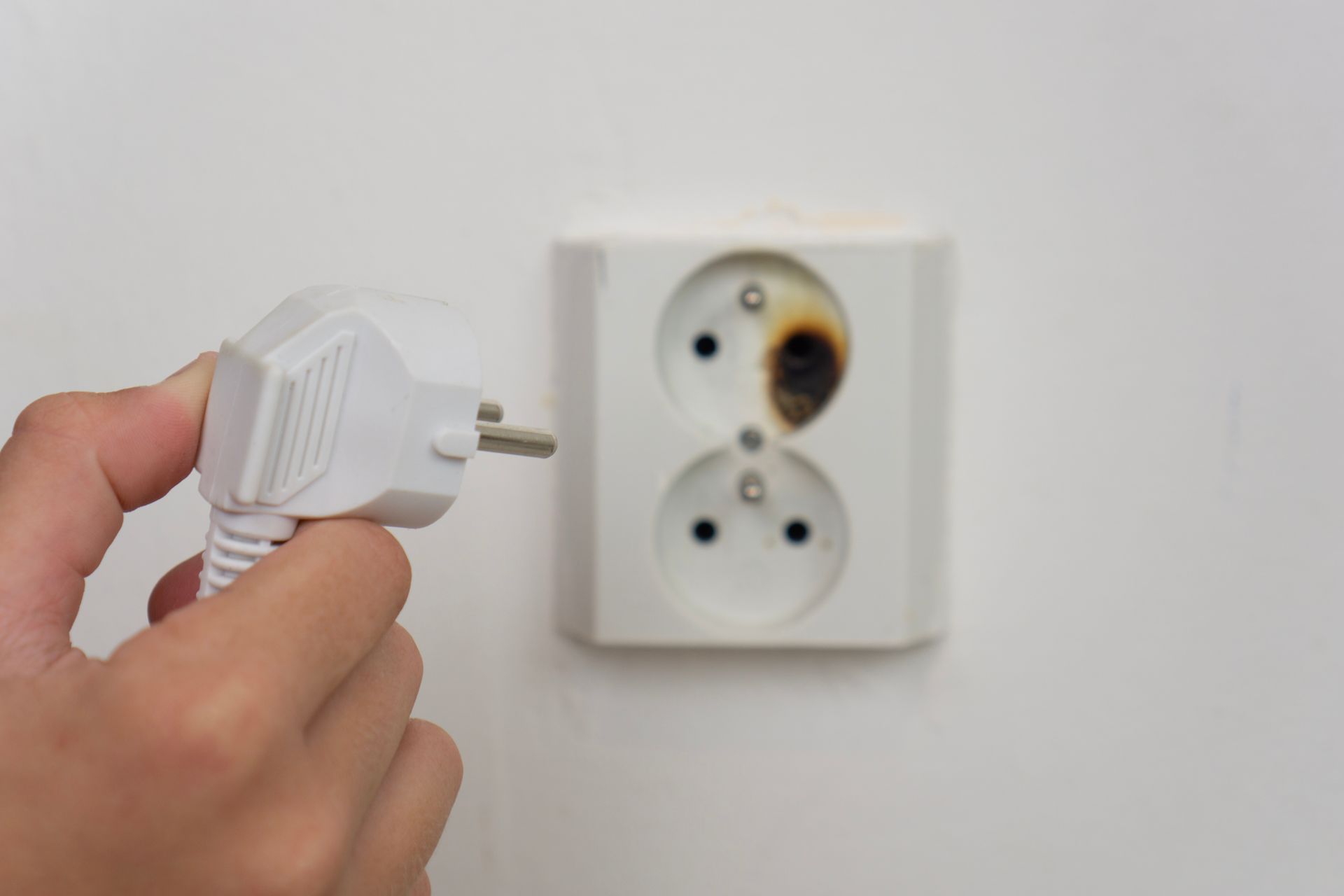A hand holds an electrical plug near a wall outlet with a charred, burnt socket.