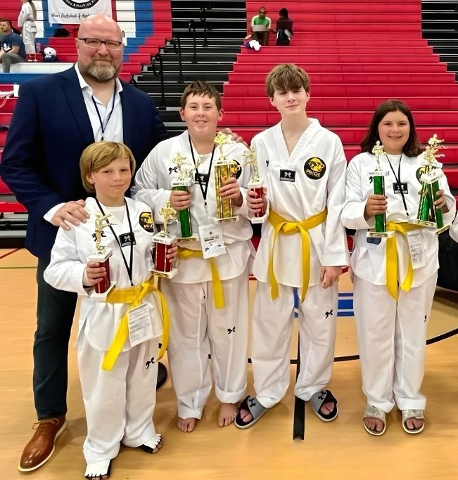 Family in Jiu-Jitsu gis poses. Indoors, blue mats, a smiling woman in the middle, around a seated man with a beard.