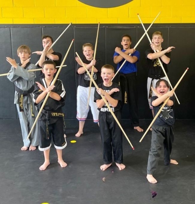 Five people in jiu-jitsu uniforms on a blue mat, including a man with a red belt, posing in a gym.