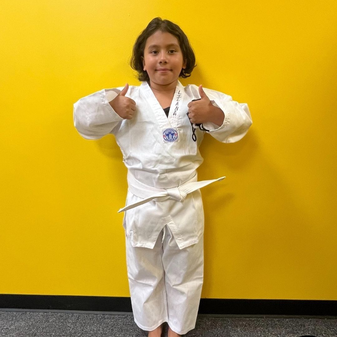 Two children practicing Brazilian Jiu-Jitsu on a mat. One child in white gi has the other in a leg lock. A girl watches.