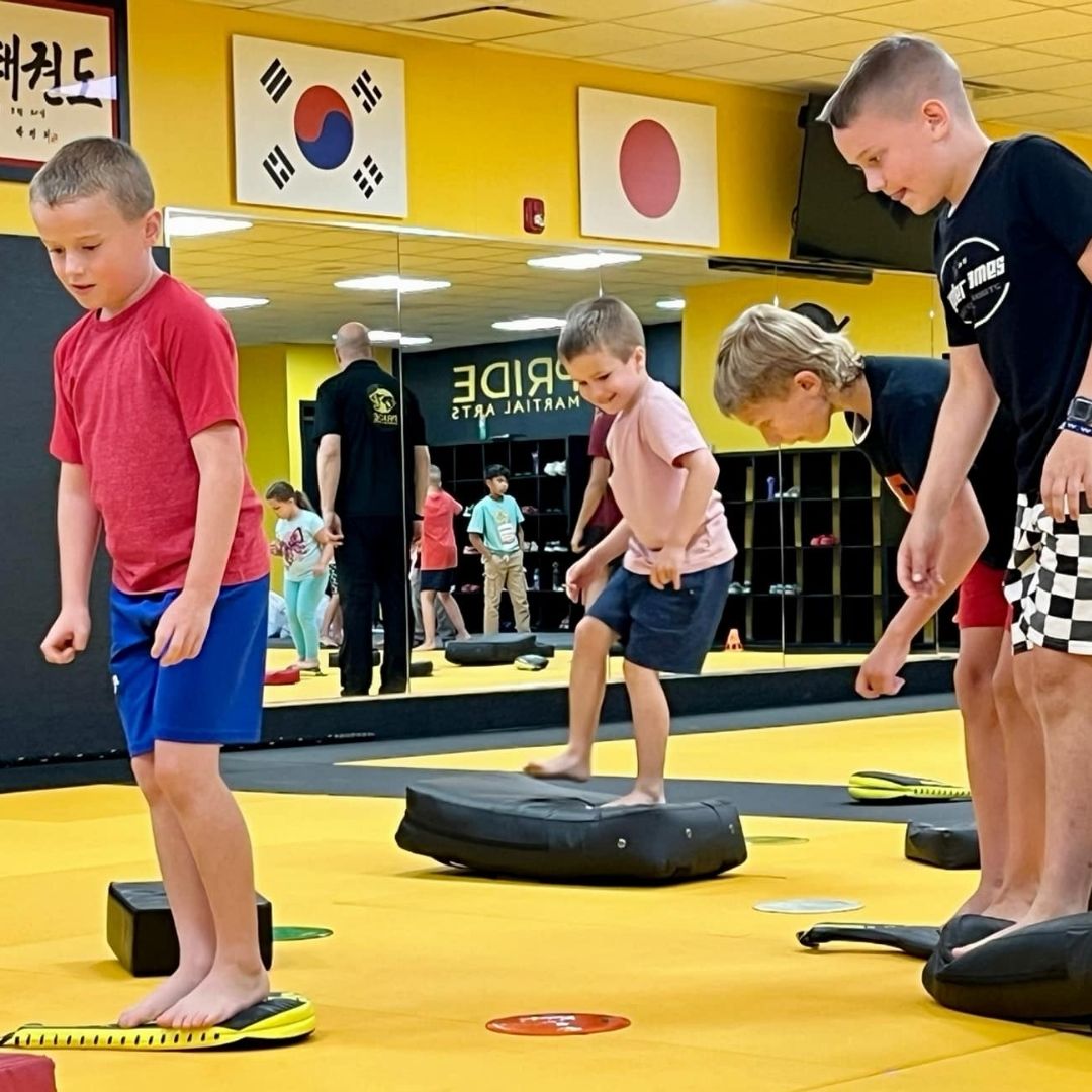 Children in colorful gis play a game on a mat. Two instructors pull a student in blue.