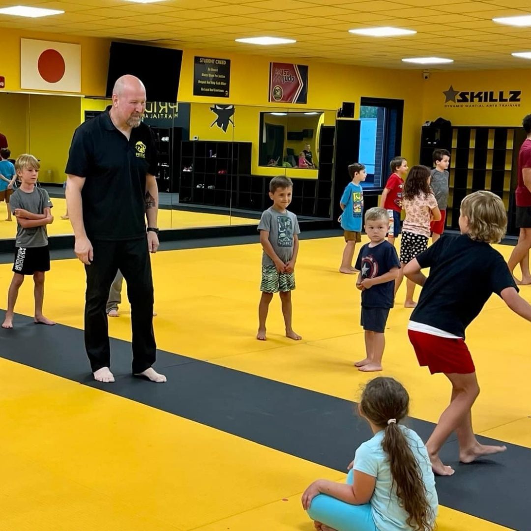 People practicing Brazilian Jiu-Jitsu. One person on the ground, another on top; two others grappling nearby. Black gis, indoor setting.