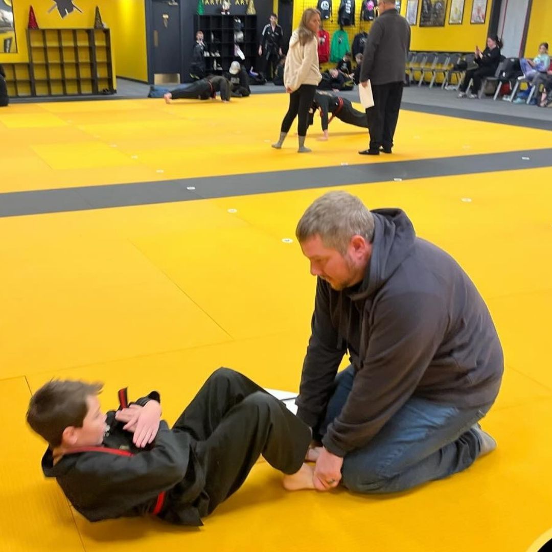 Two young children in blue and black martial arts uniforms grappling on a gym floor, one smiling.