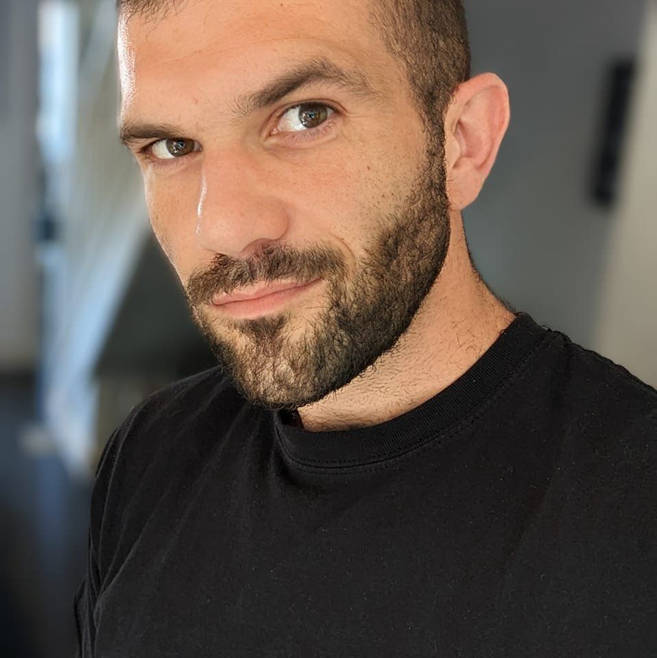 Man with short dark hair and beard in a black shirt; looking at the camera, indoors.