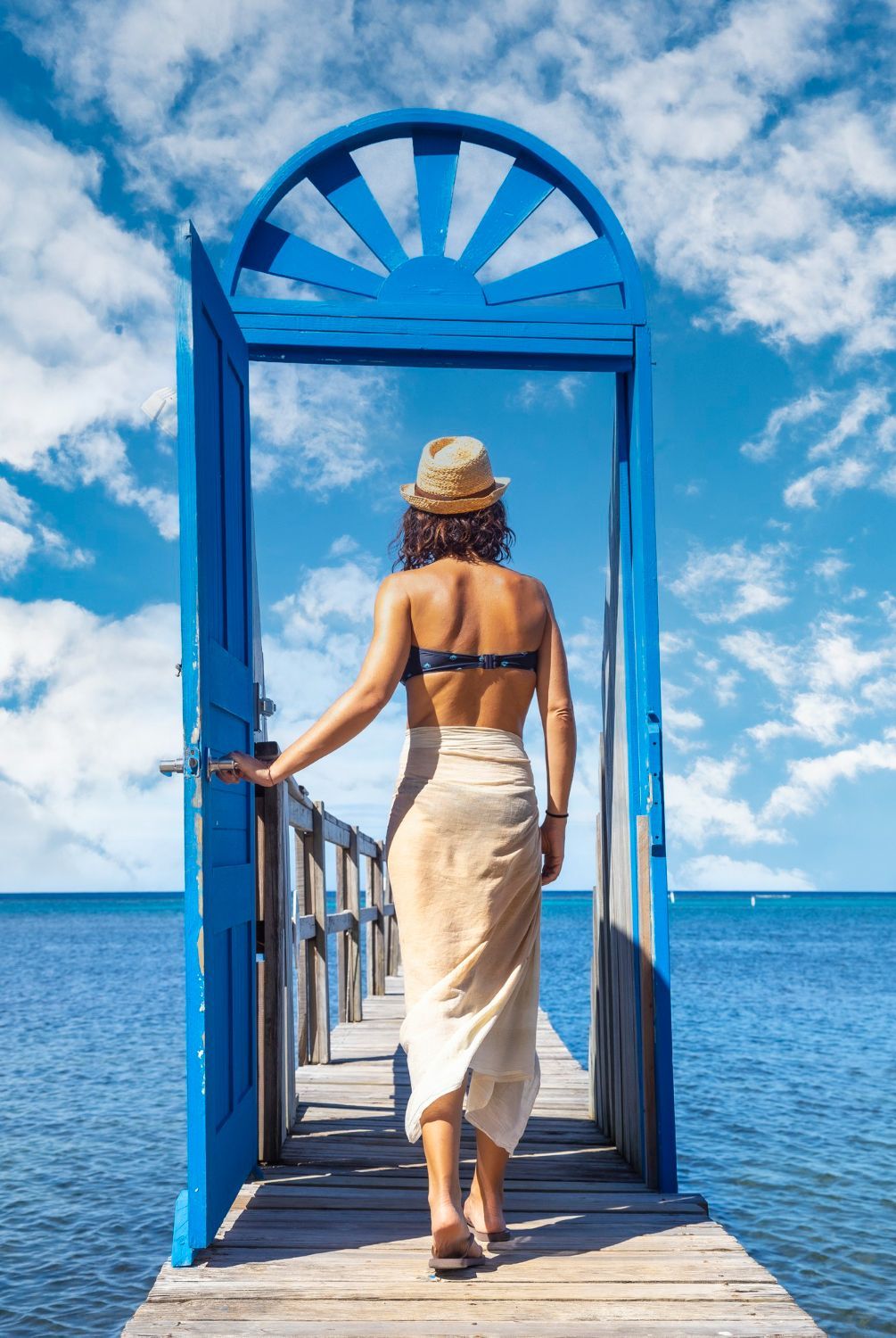 Woman in swimsuit and hat walks towards blue arched doorway on a pier over the ocean.