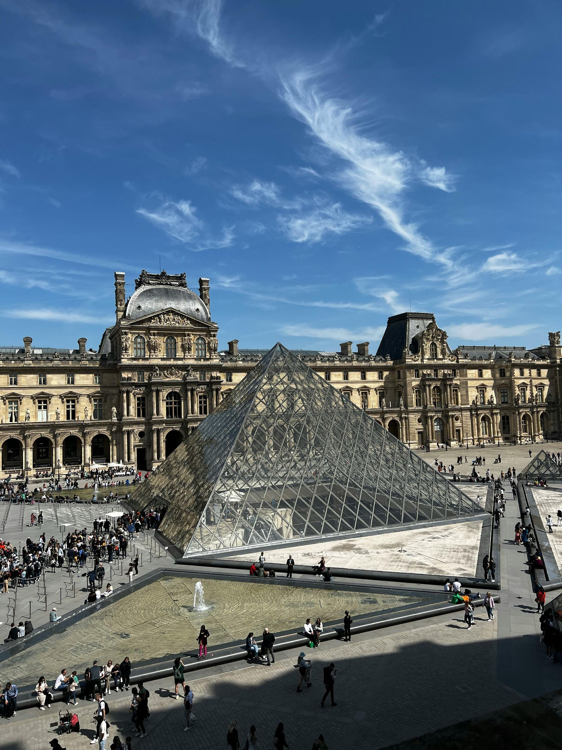 Louvre Museum with glass pyramid in Paris, France; people gathered outside on a sunny day.