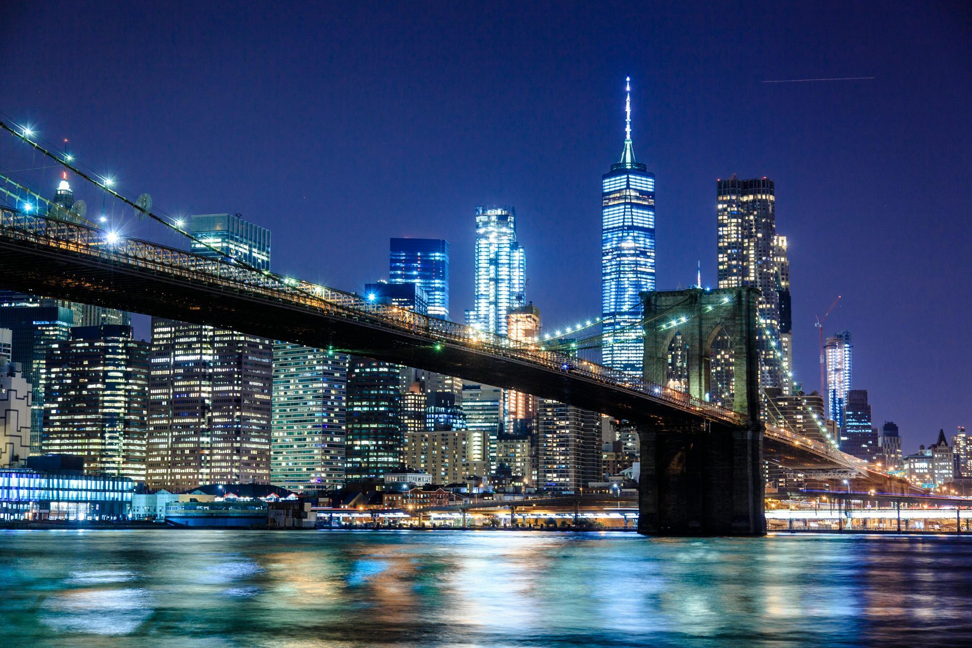 Brooklyn Bridge at night with city skyline, illuminated skyscrapers, and water reflections.