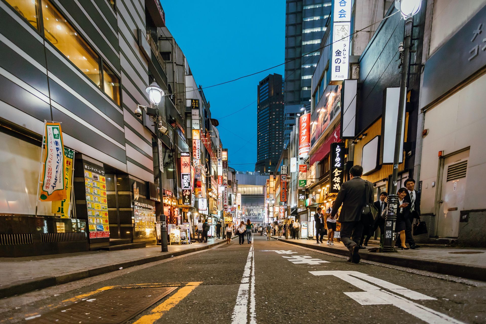 Narrow street in a city at dusk, lined with buildings, signs, and people walking.