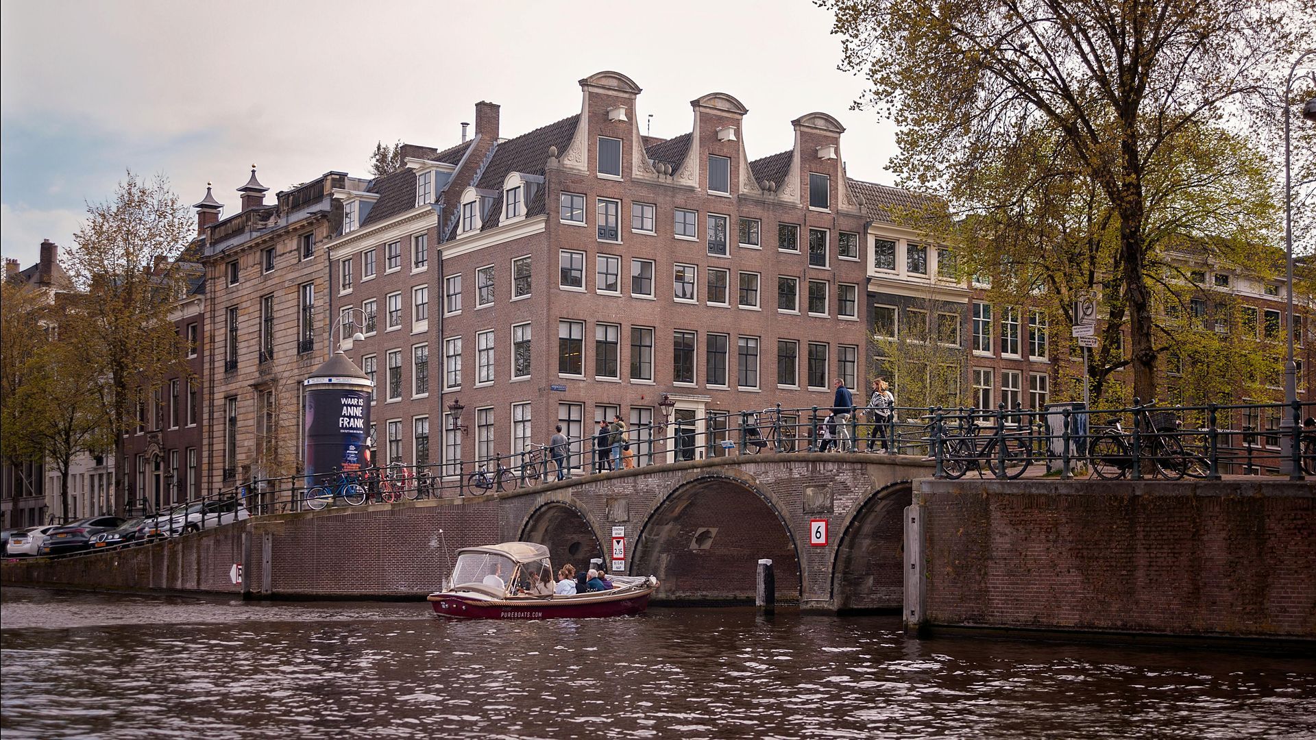 Canal with brick buildings, arched bridge, boat, and people in Amsterdam.