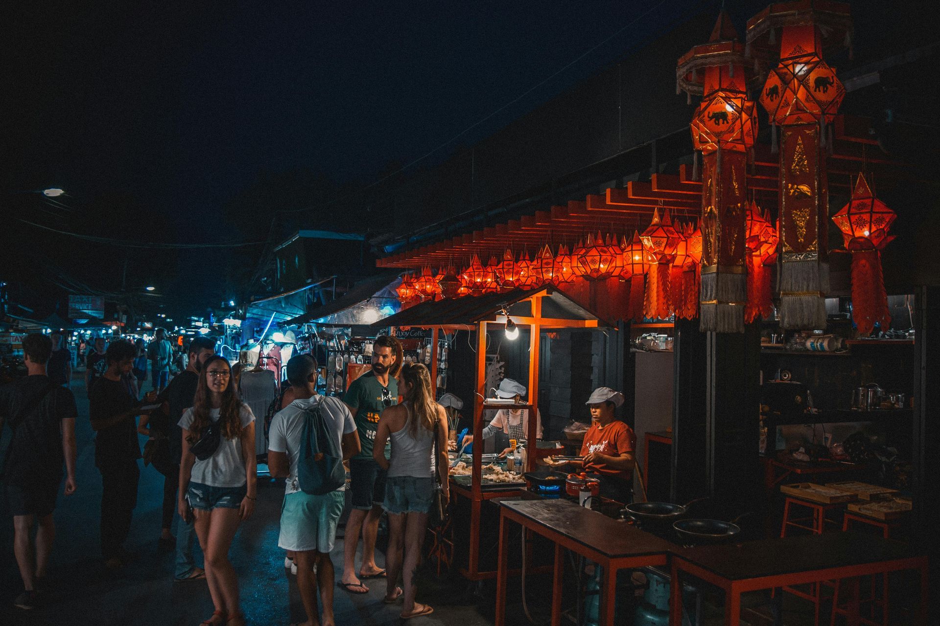 Night market scene: people walking, food stalls lit by red lanterns, nighttime.