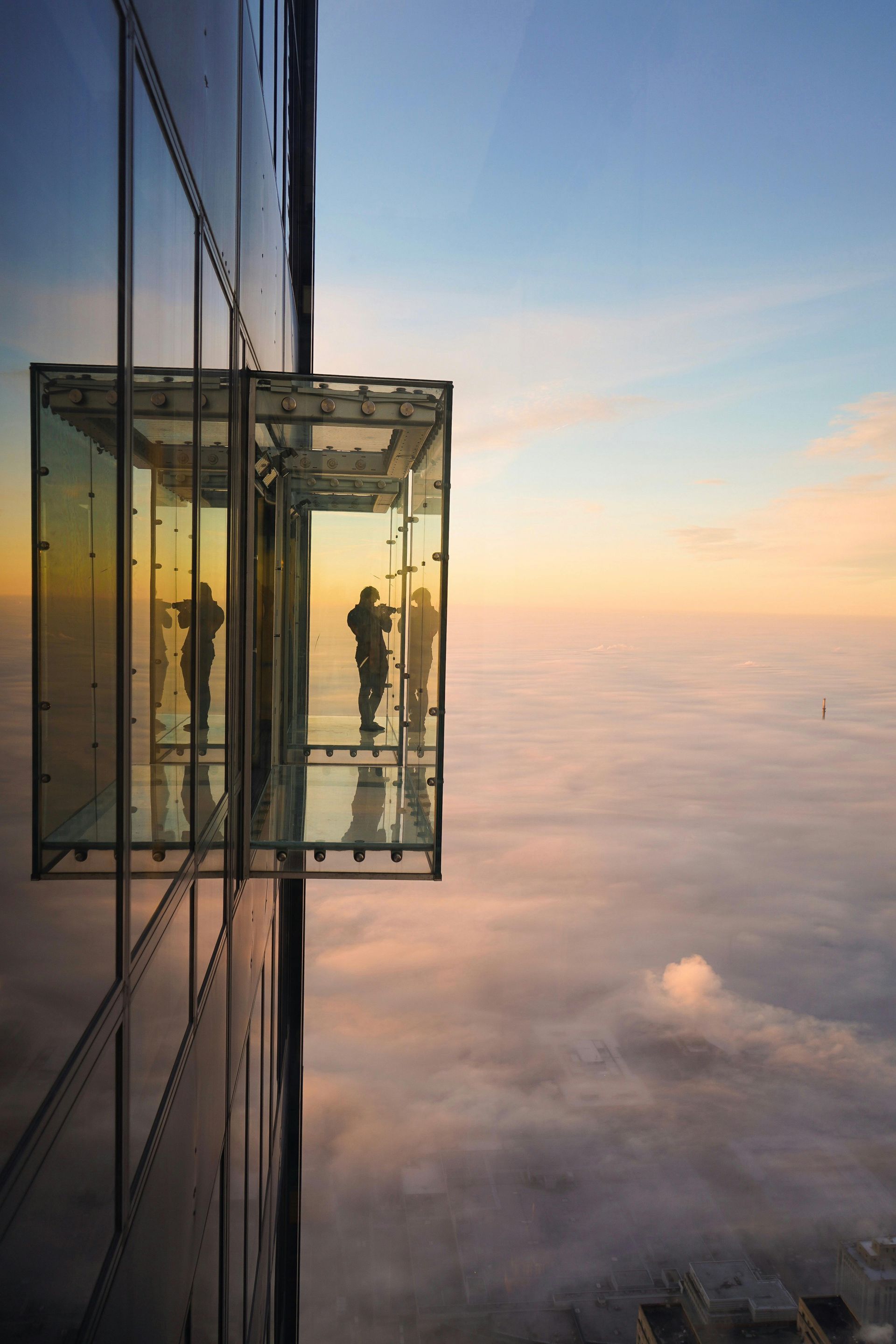 People in a glass box extending from a skyscraper, overlooking a sea of clouds at sunset.