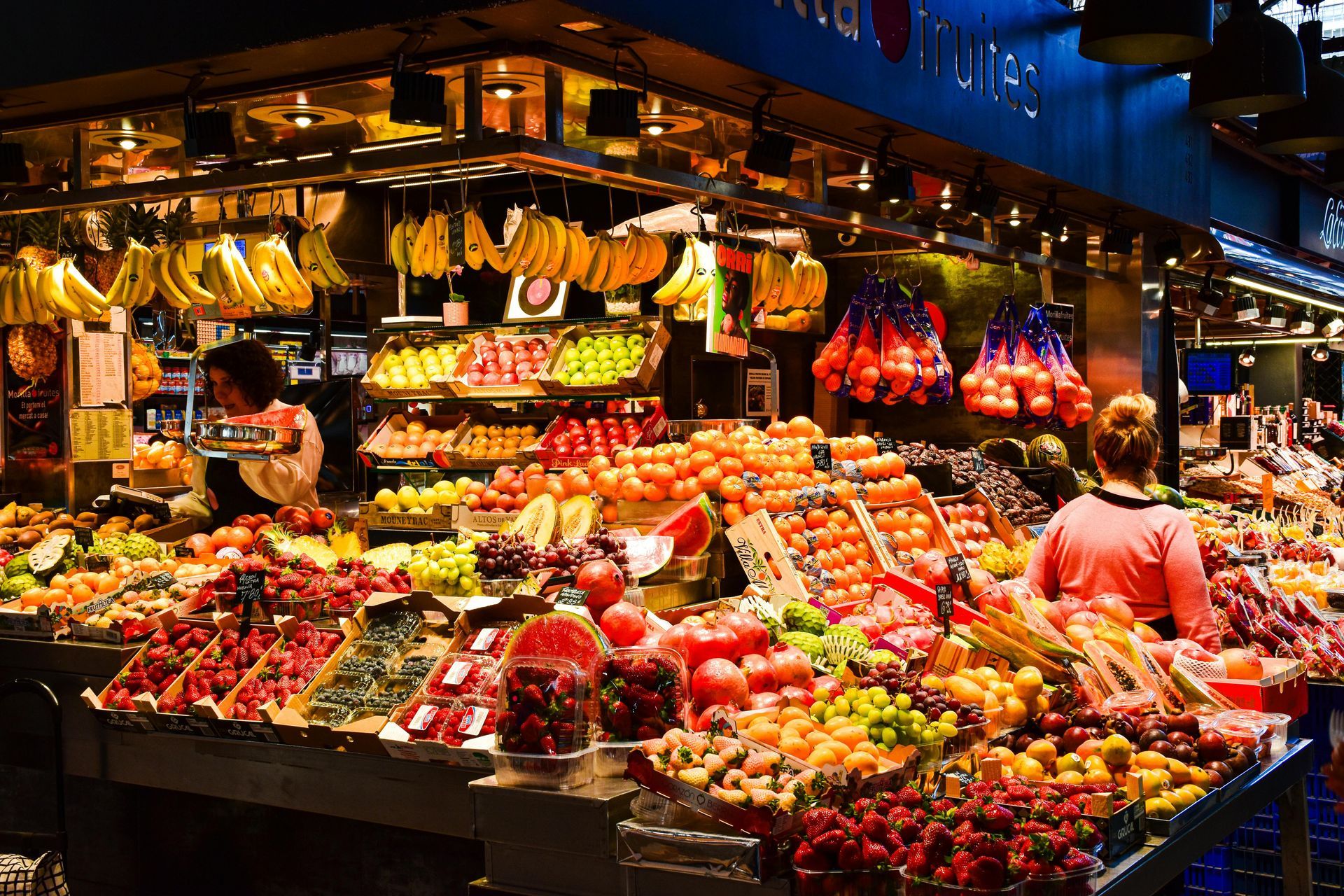 Fruit market stall with bananas, grapes, tomatoes, and vendors.