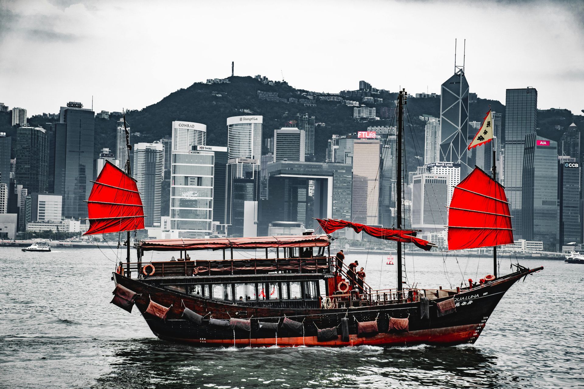 A traditional red-sailed junk boat sailing in Hong Kong harbor with city skyline in the background.
