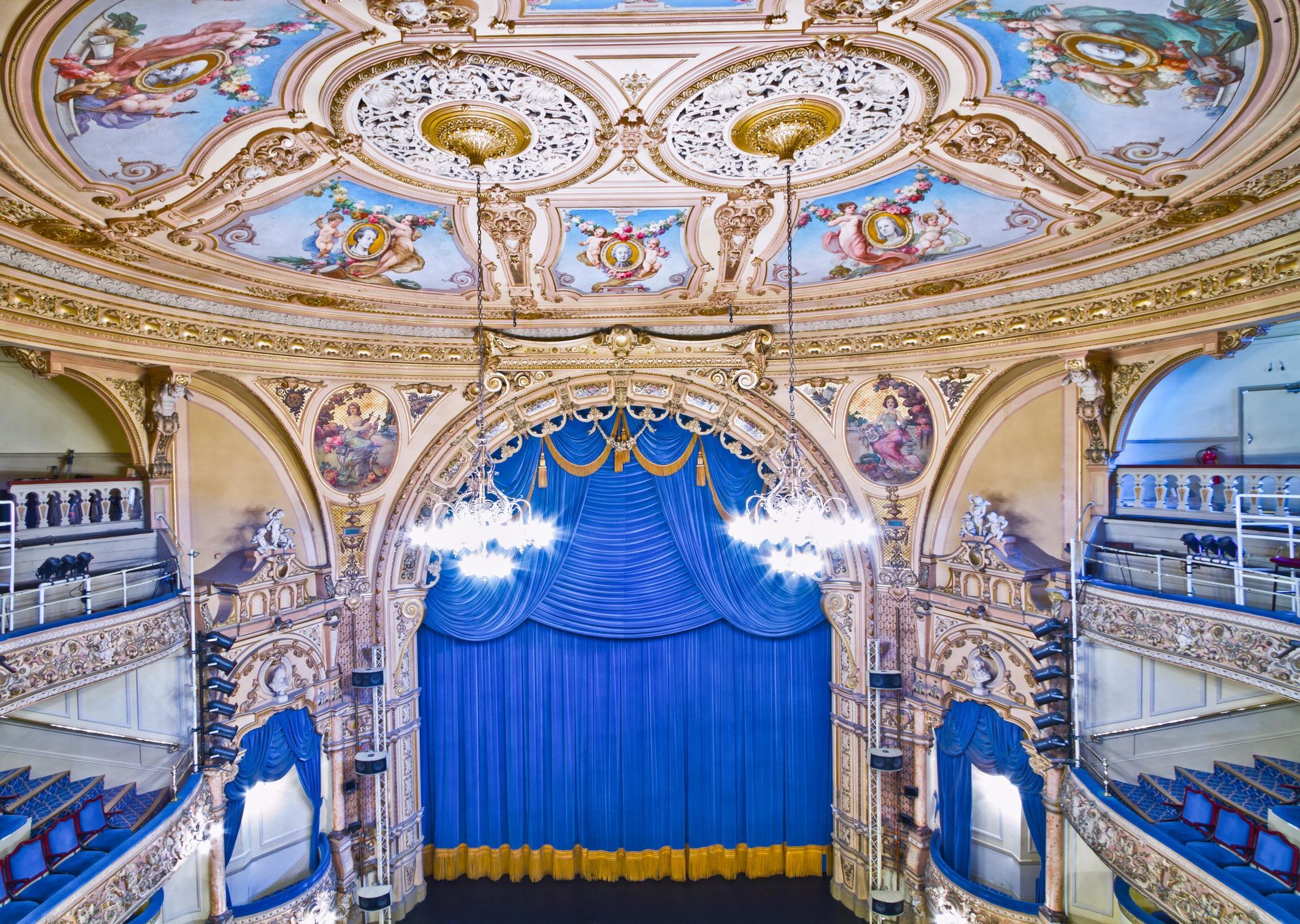 Ornate theater interior with blue curtain, gold detailing, and painted ceiling.