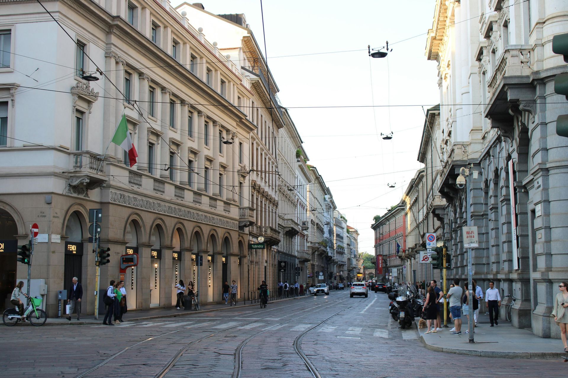 Street view in Milan, Italy, with buildings, pedestrians, cars, and a green Italian flag.
