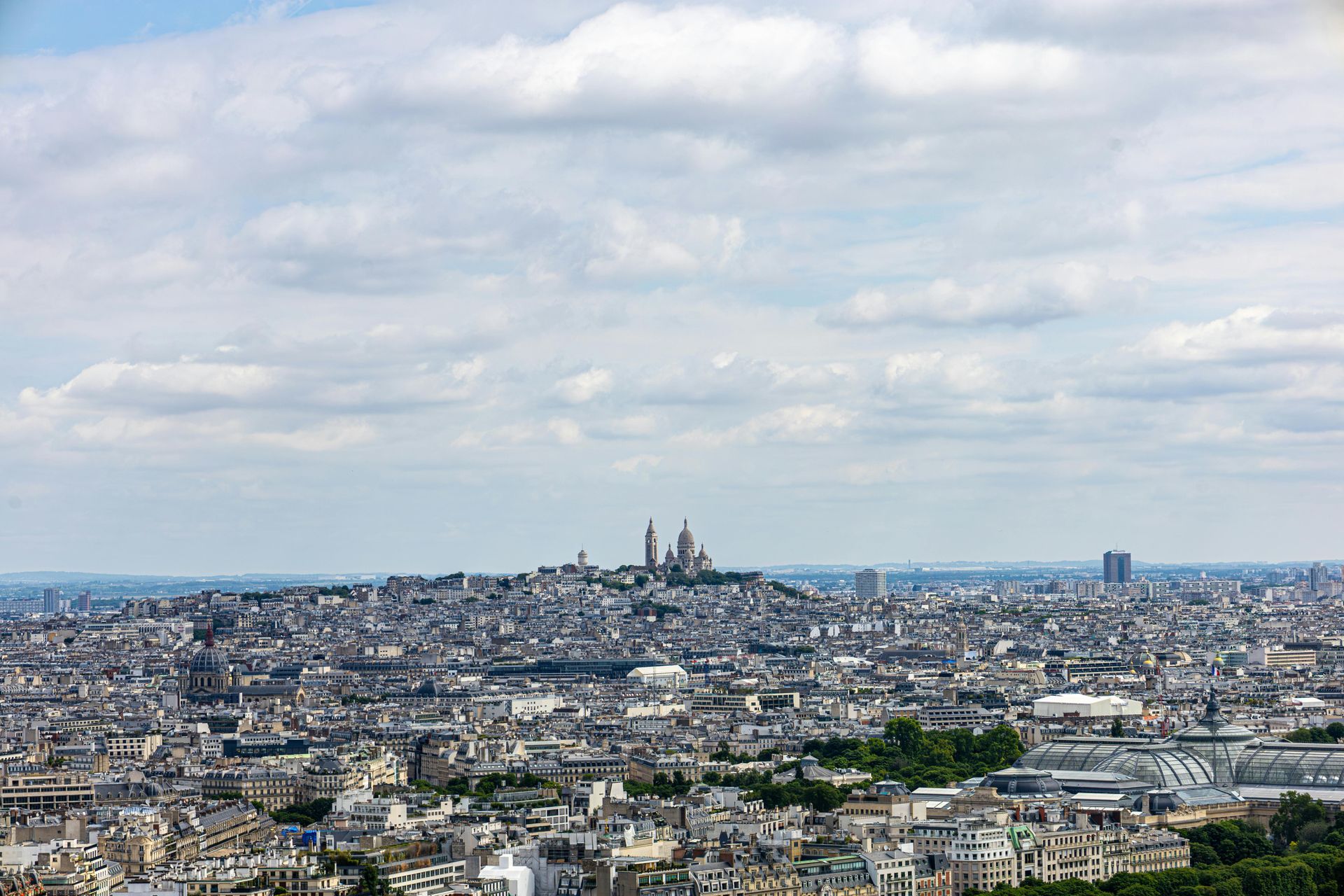 Panoramic view of Paris with the Sacré-Cœur Basilica on a hill under a cloudy sky.