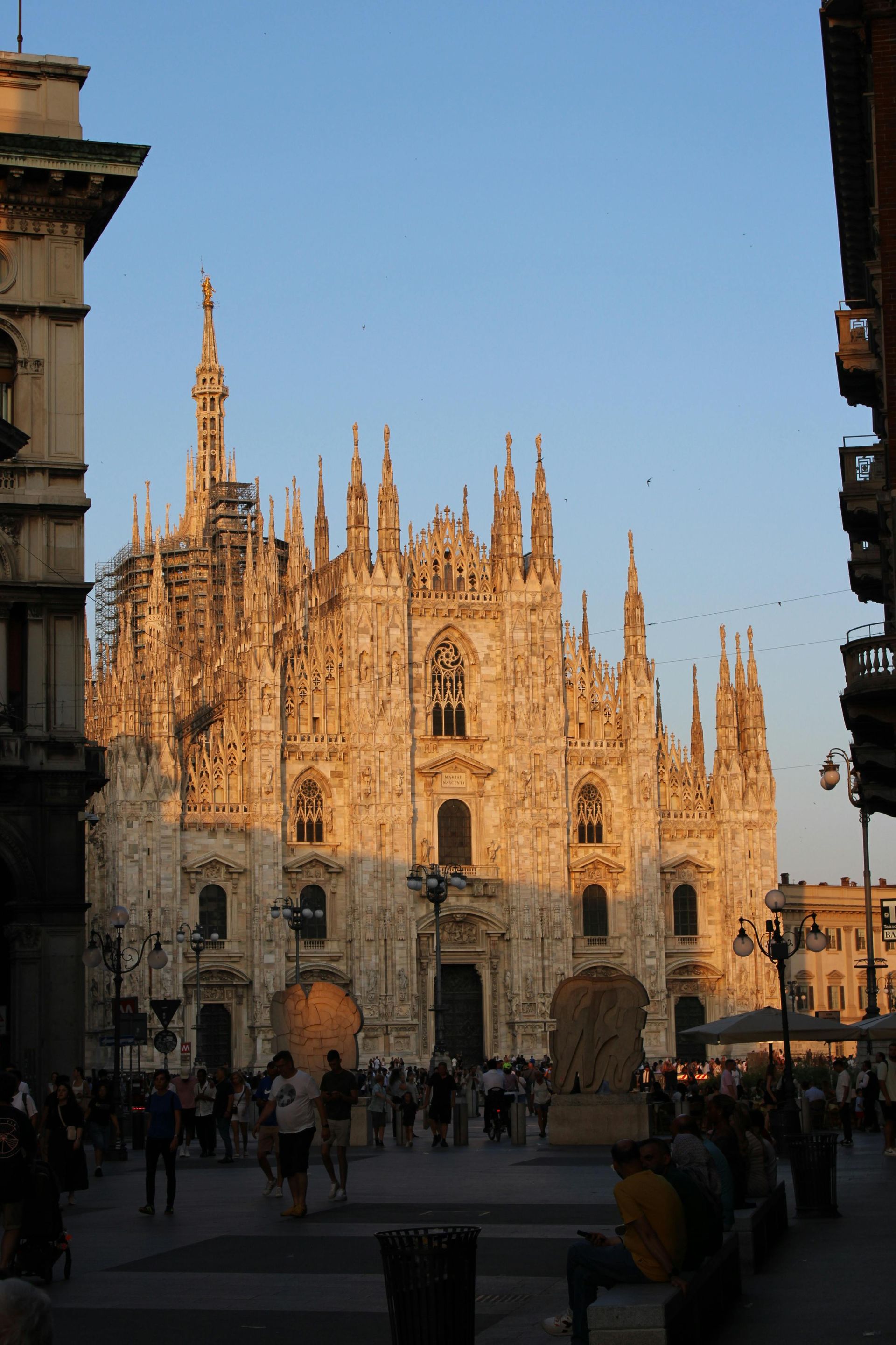 Milan Cathedral bathed in warm sunlight, with people milling about in the square.