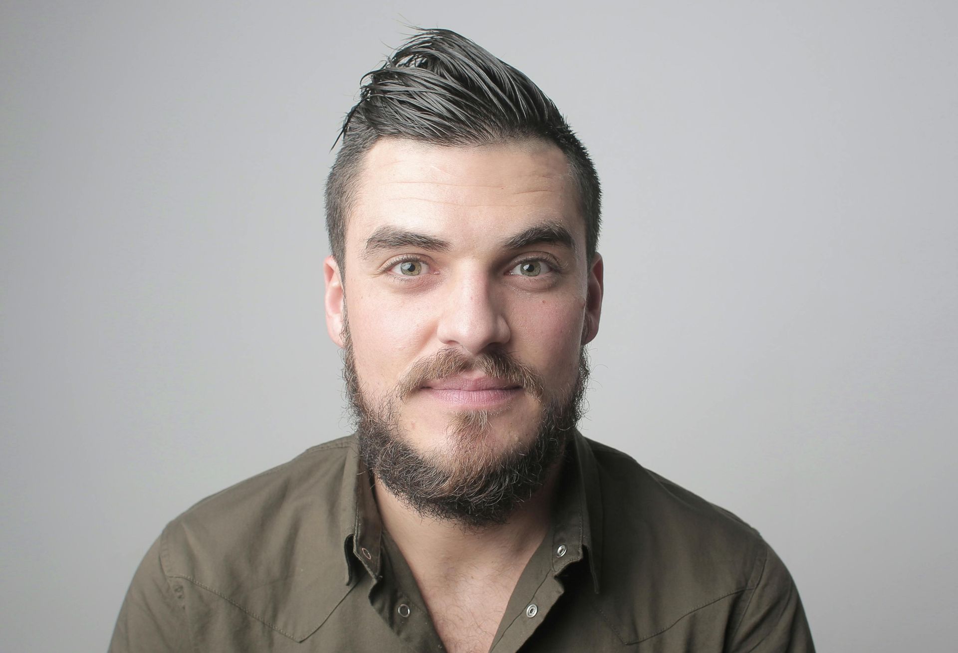 Man with a styled, dark haircut, and beard in a green shirt against a gray backdrop, smiling.
